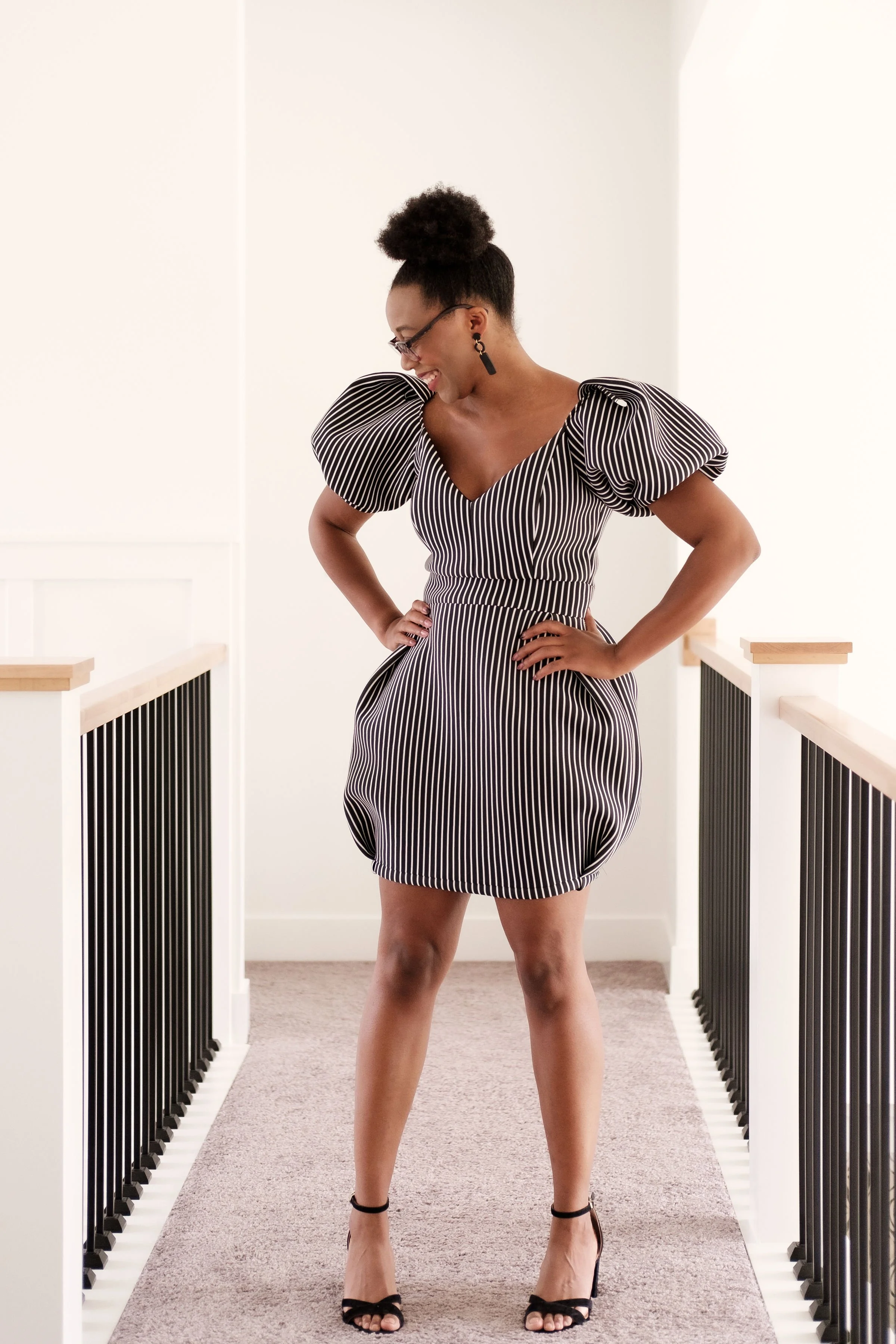 A woman in a black and white striped dress with puff sleeves stands on a carpeted hallway, smiling and looking down, with hands on hips.