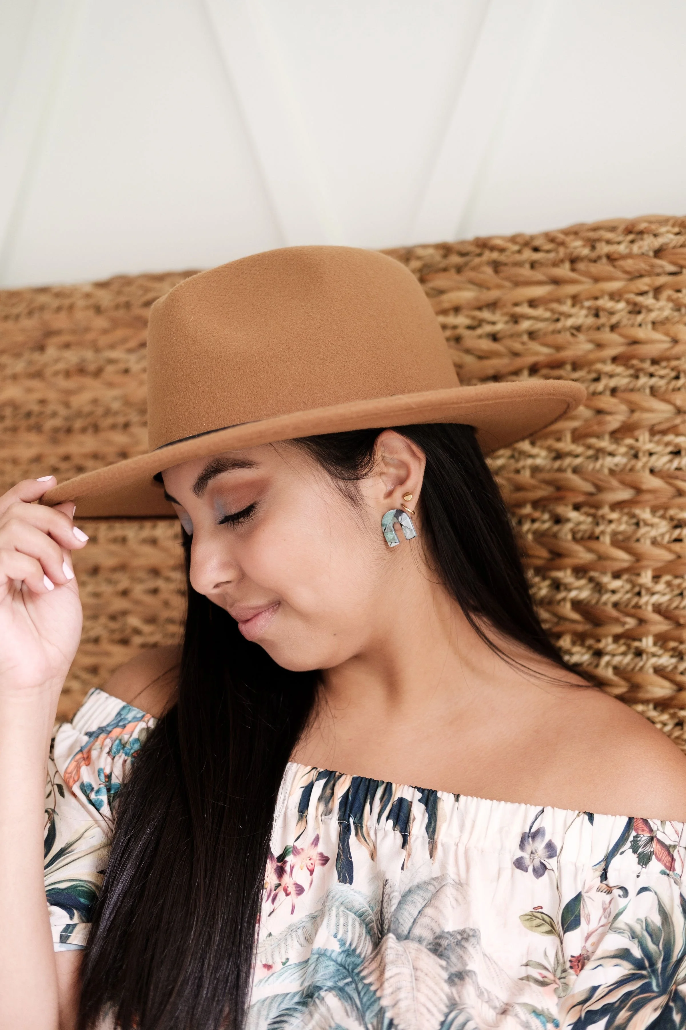 A woman with long dark hair, closed eyes, and a gentle smile, wearing a wide-brimmed brown hat, floral off-shoulder top, and earrings, sitting against a woven headboard.