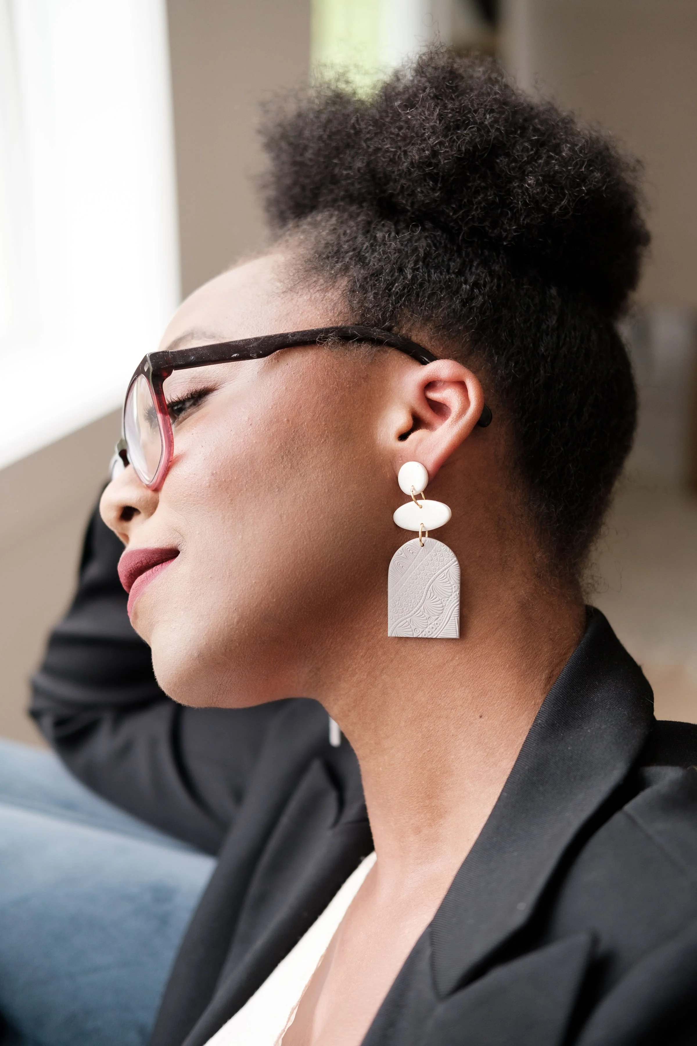 Close-up profile of an elegant woman with curly hair styled in a bun, wearing glasses, large statement earrings, and a black blazer, sitting by a window.