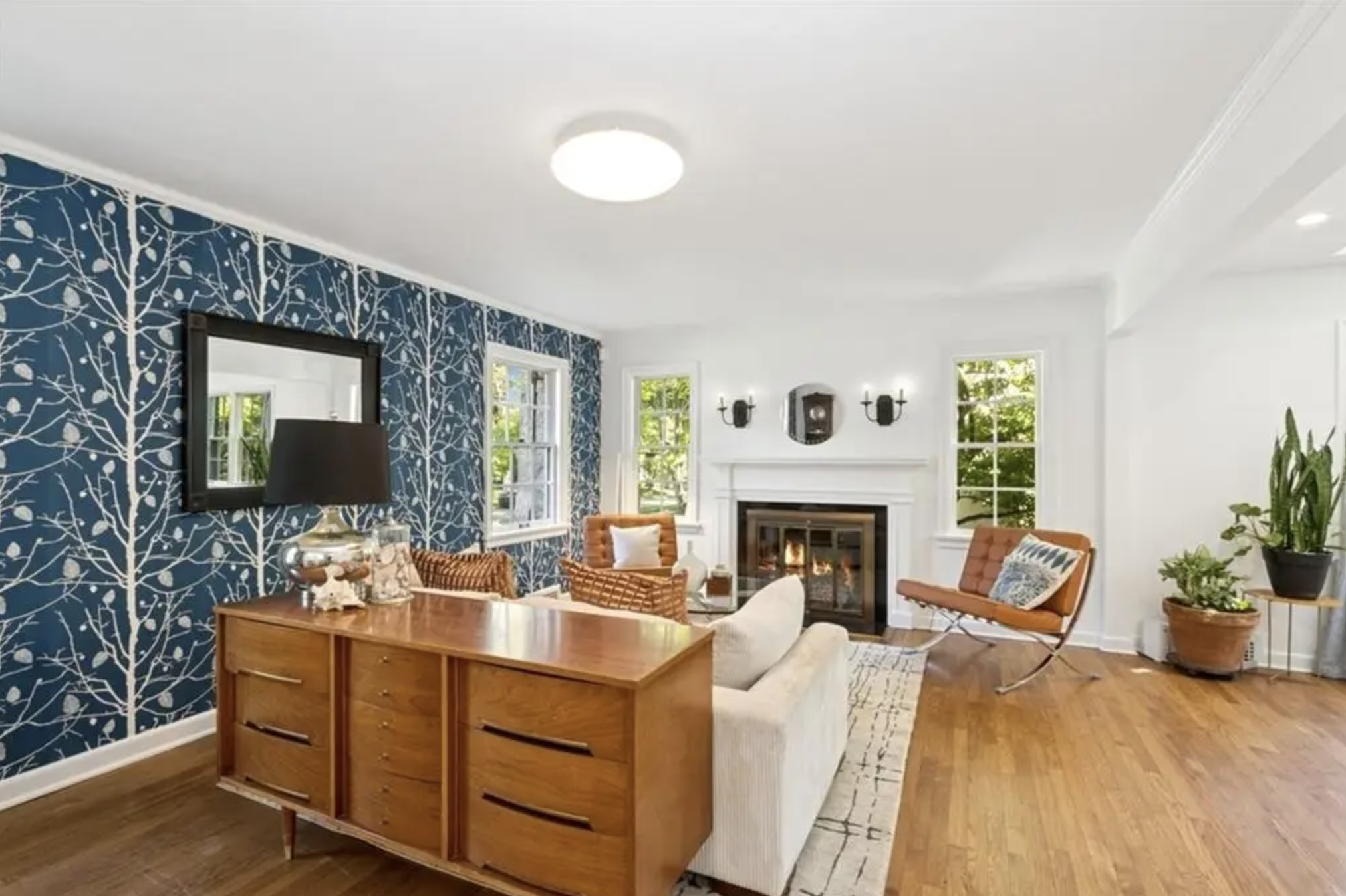 Living room with white walls, hardwood floors, a fireplace, a beige sofa, a wooden sideboard, an orange chair, a bench, and large windows. One wall has blue wallpaper with white tree patterns.