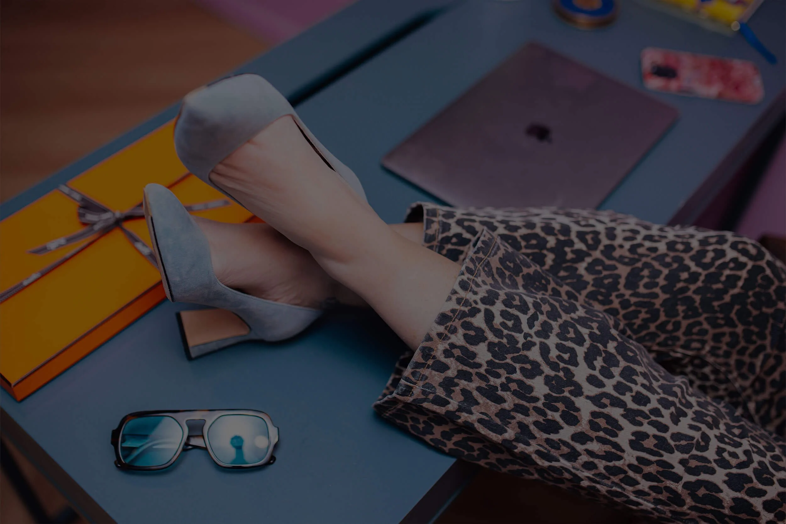 A person with leopard print pants and grey high heels sitting at a desk with folded legs on it. On the desk are sunglasses, an orange box, a purple laptop, a smartphone, and a patterned phone case.
