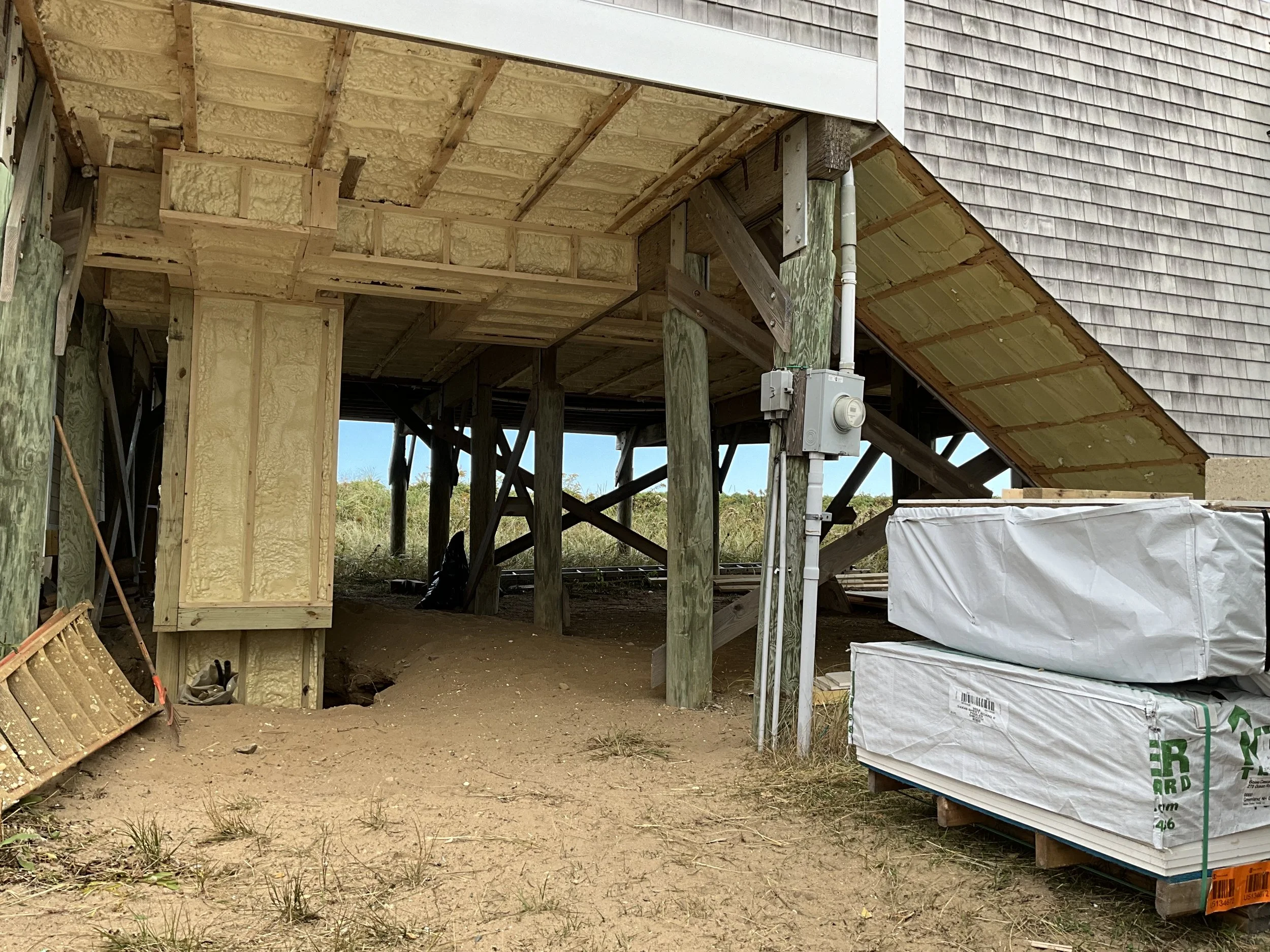 Under elevated construction, wooden frame building with insulation installed, construction materials stored nearby, and outdoor environment visible.