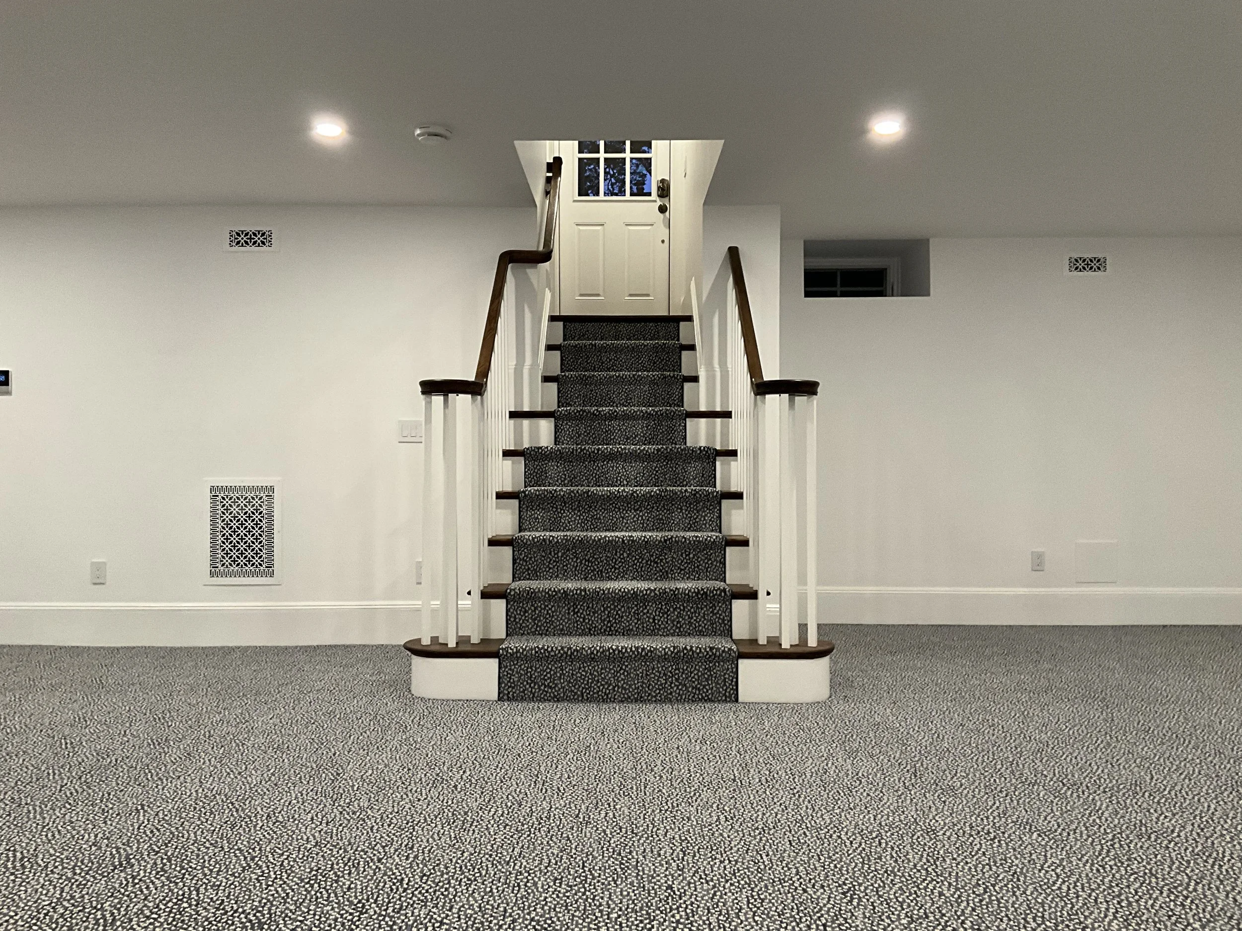 Empty carpeted staircase leading to a white door at the top in a plain white room with recessed ceiling lights.