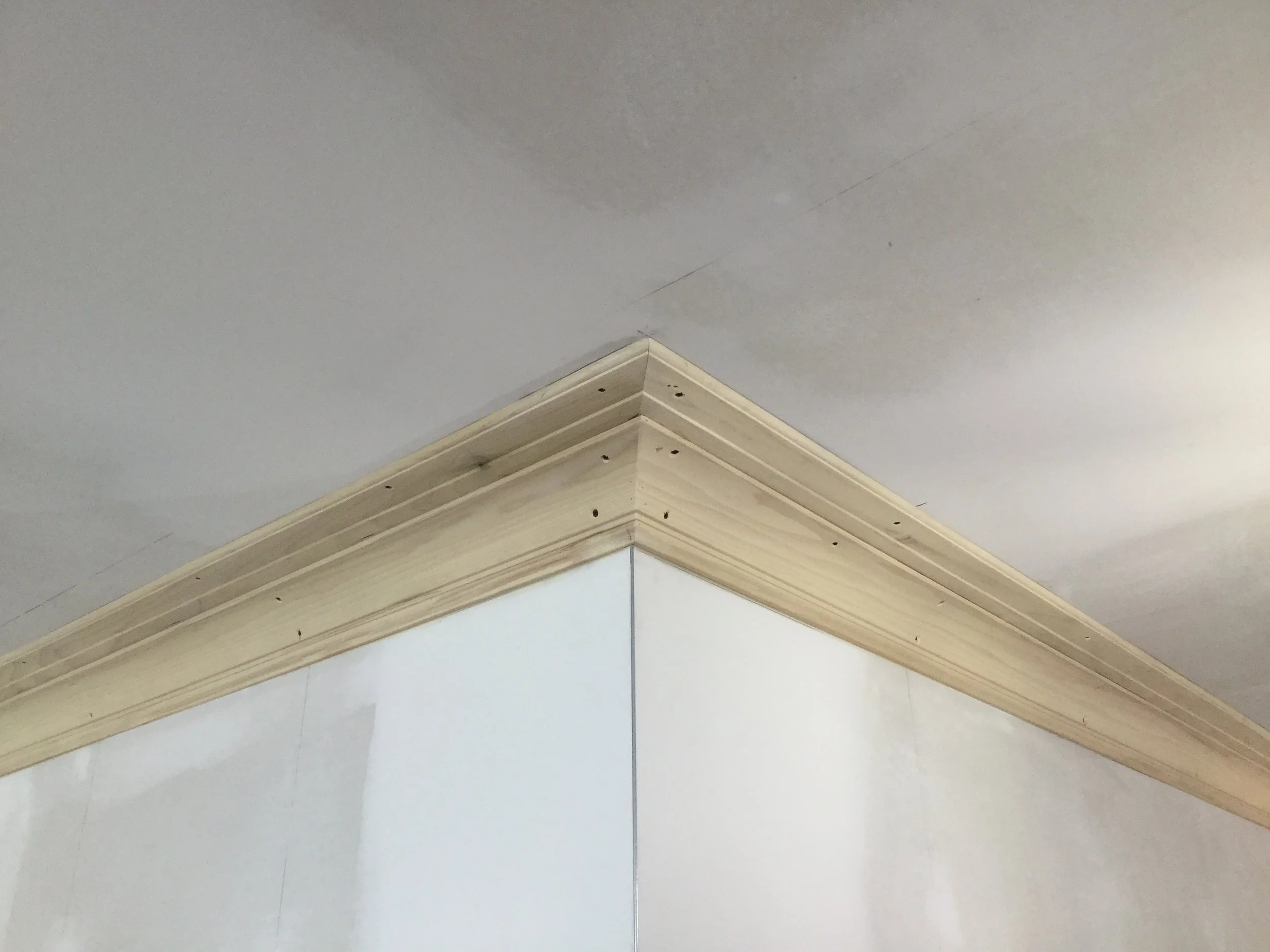 Corner of a room showcasing crown molding detail on the ceiling and wall, with visible nail holes in the wood trim.