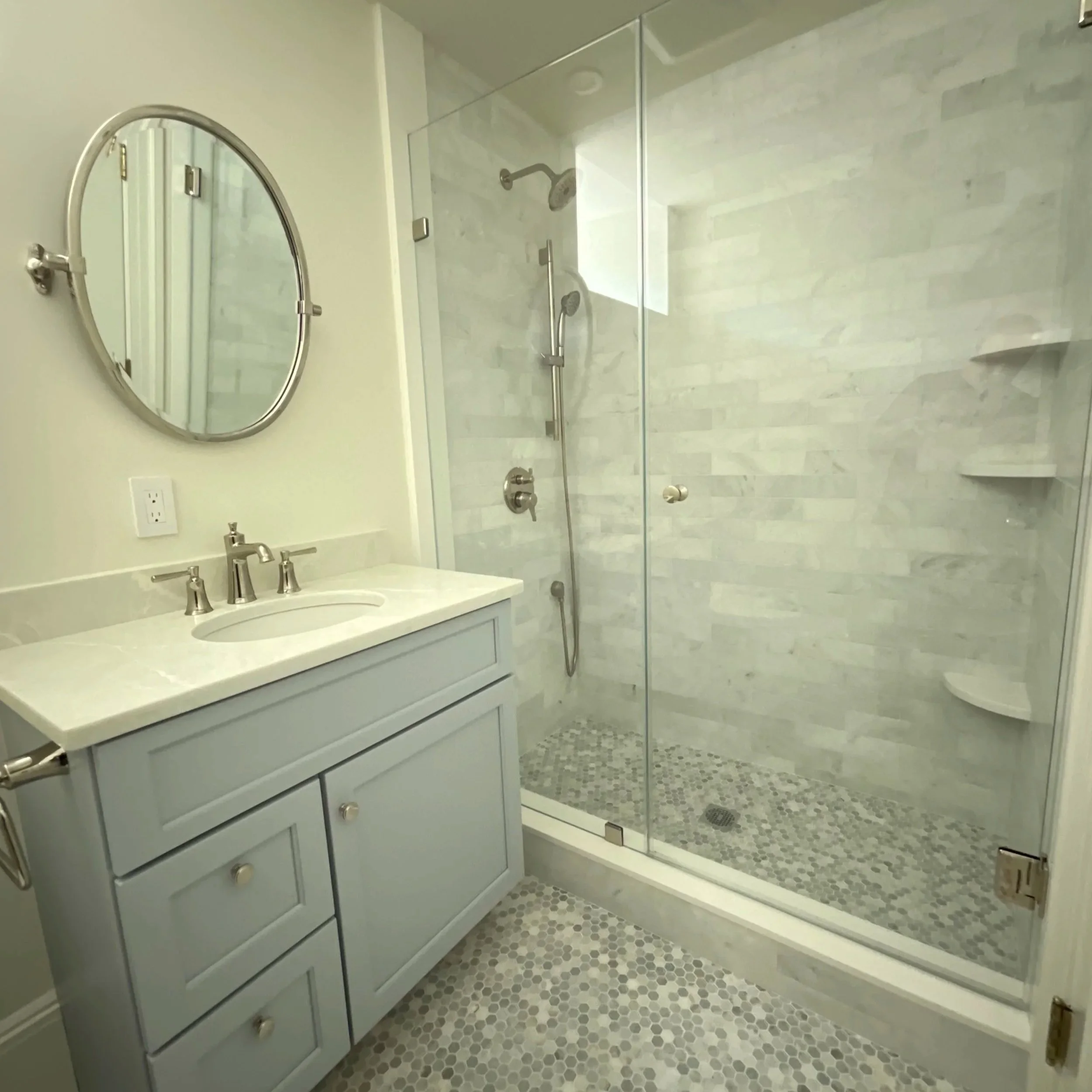 Bathroom with a light gray vanity sink, mirror, and a glass-enclosed shower with tiled walls and pebble tile floor.