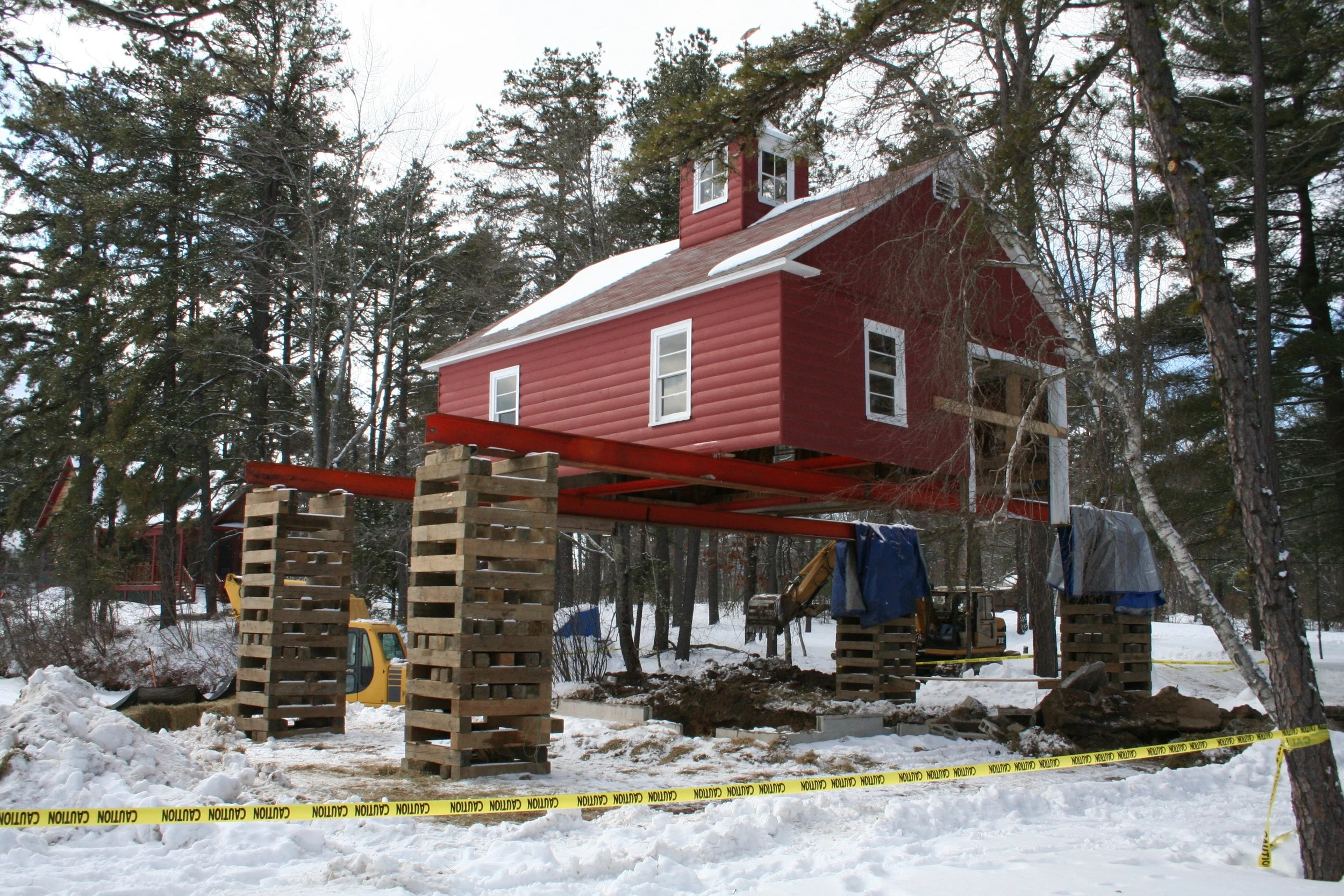 Construction site with a house on stilts surrounded by snow. The house is painted red with white trim and is elevated on large wooden pallets, with construction equipment and caution tape in the foreground.