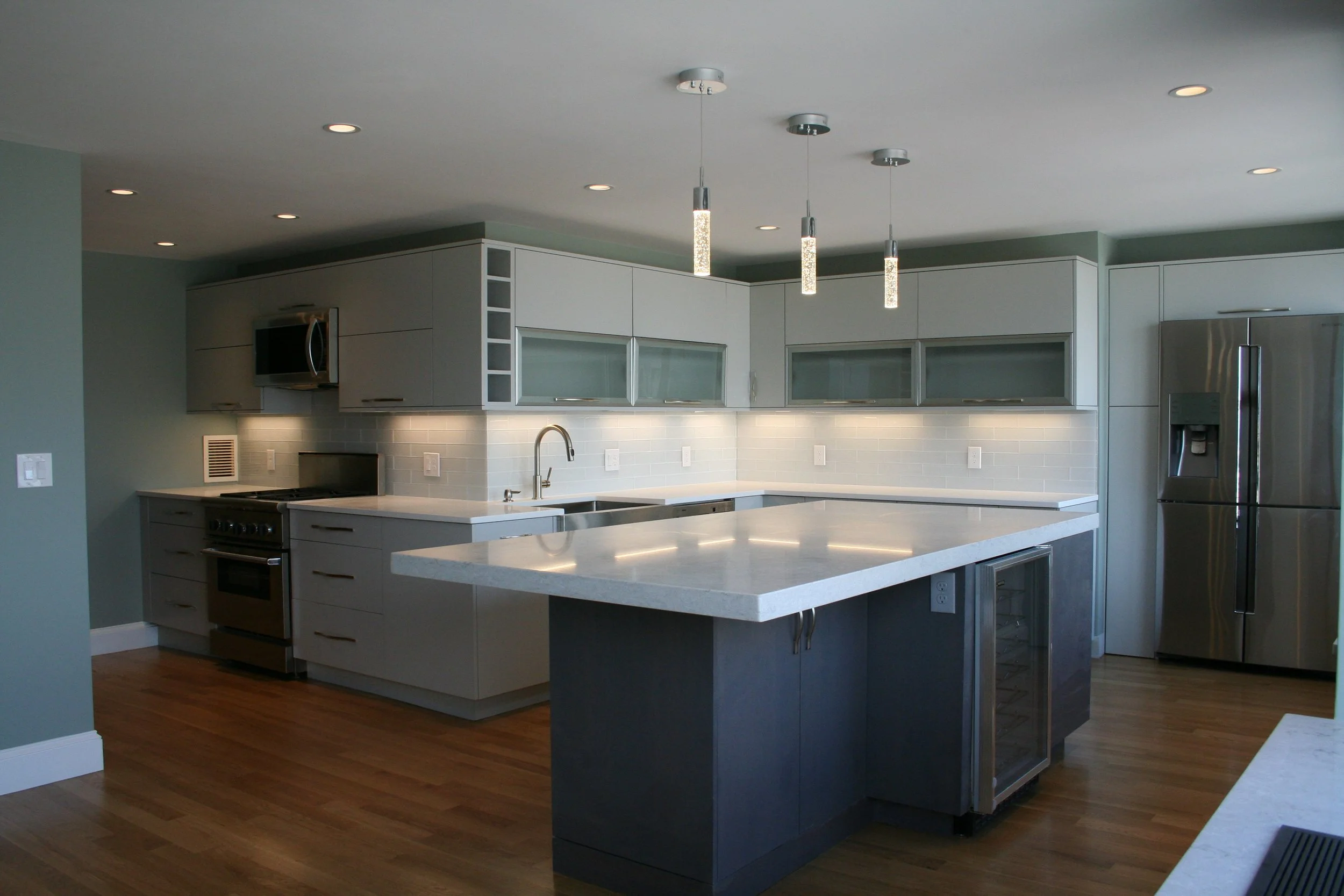 Modern kitchen with white and gray cabinets, stainless steel appliances, wooden floors, an island with a wine cooler, and pendant lights.