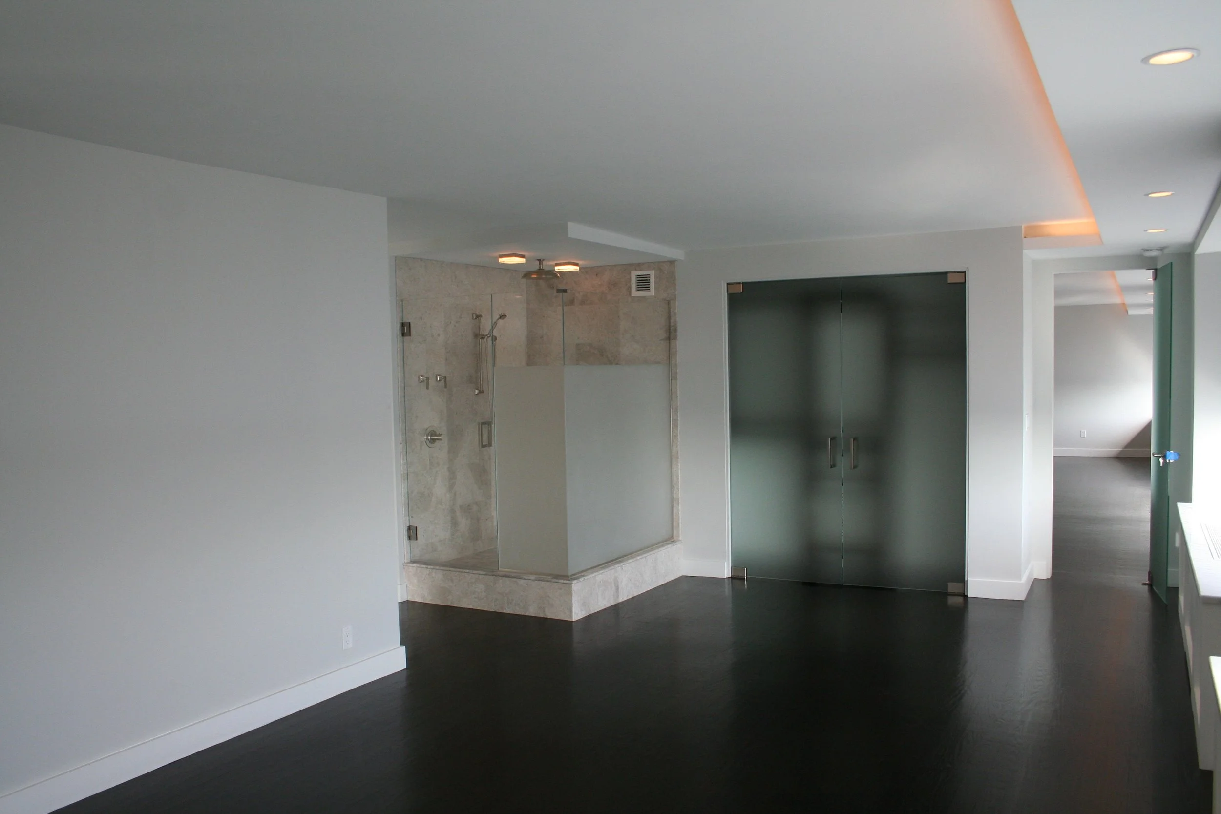 Modern interior space with a corner shower featuring glass walls, a dark wood floor, frosted glass double doors, and a room with a sloped ceiling in the background.