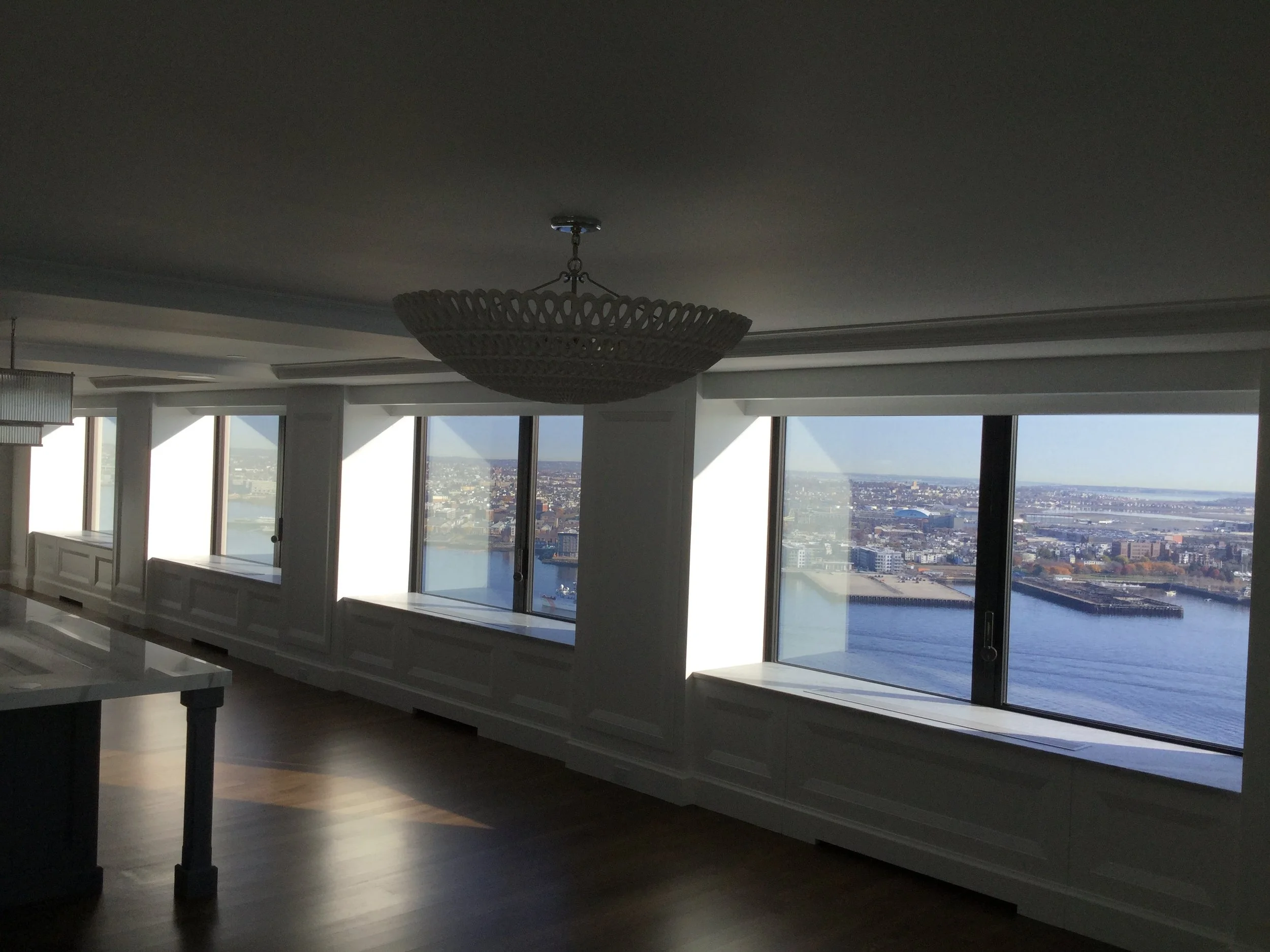 Interior view of a high-rise room with large windows showing a river and cityscape outside, pendant chandelier hanging from the ceiling, and hardwood floors.