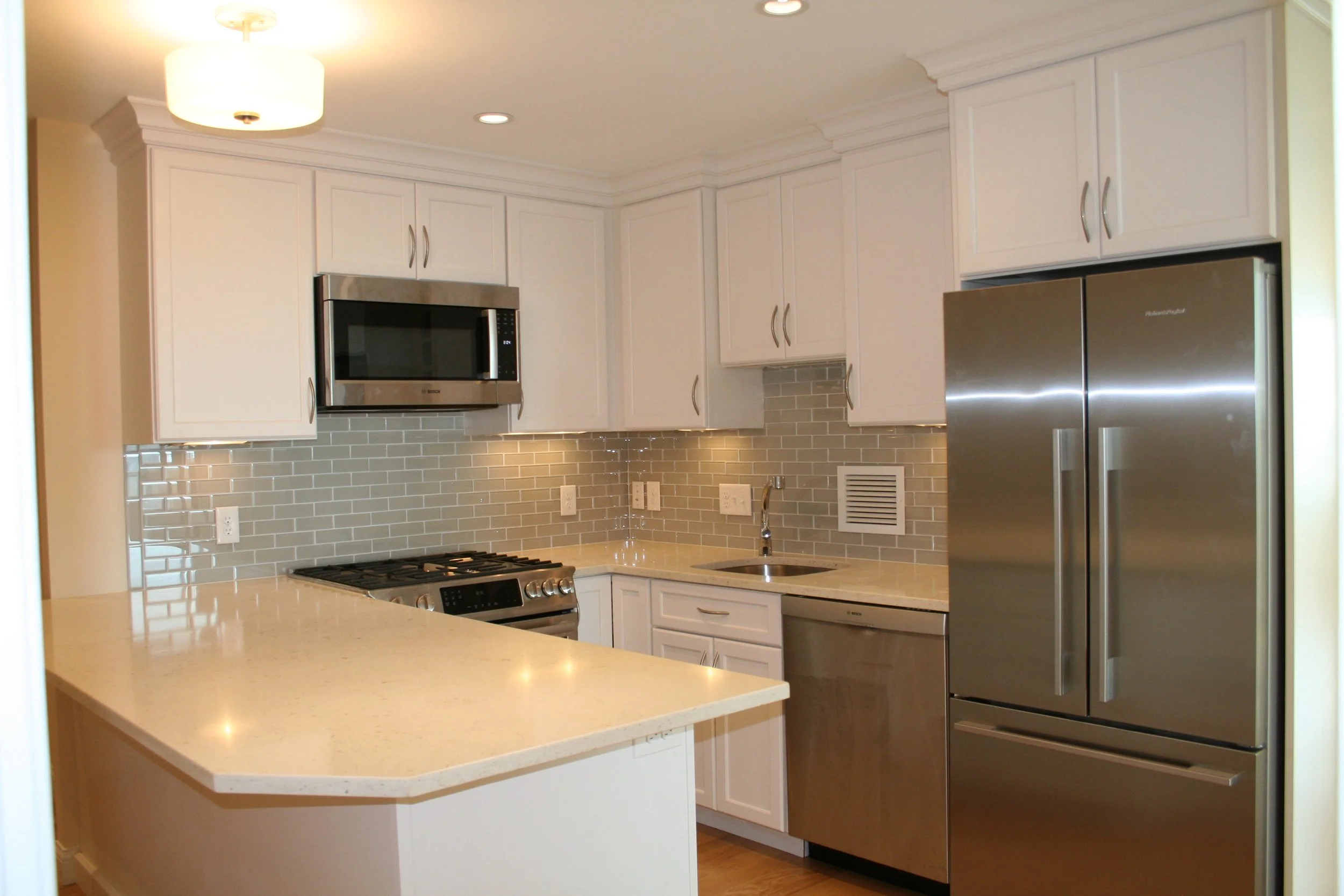 Modern kitchen with white cabinets, beige countertops, stainless steel refrigerator, dishwasher, microwave, and oven, gray subway tile backsplash, ceiling light fixture, and small window.