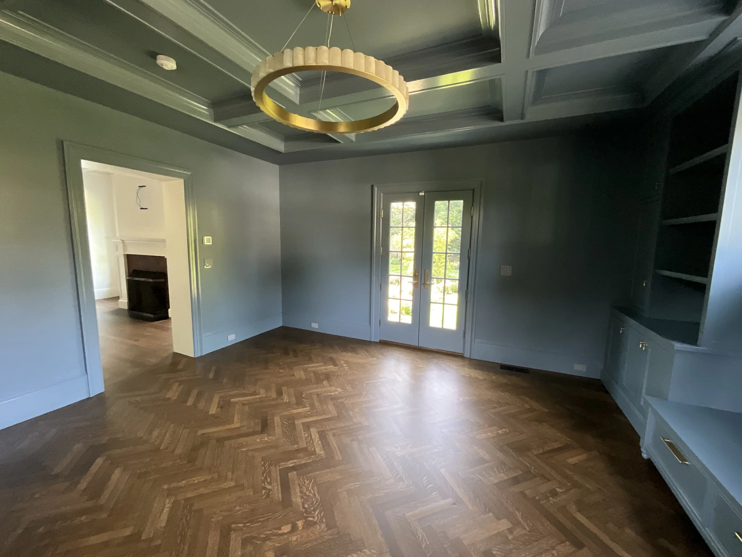 Empty living room with hardwood floors, blue walls, built-in blue shelves, and a glass double door leading outside. There is a circular chandelier hanging from the ceiling.