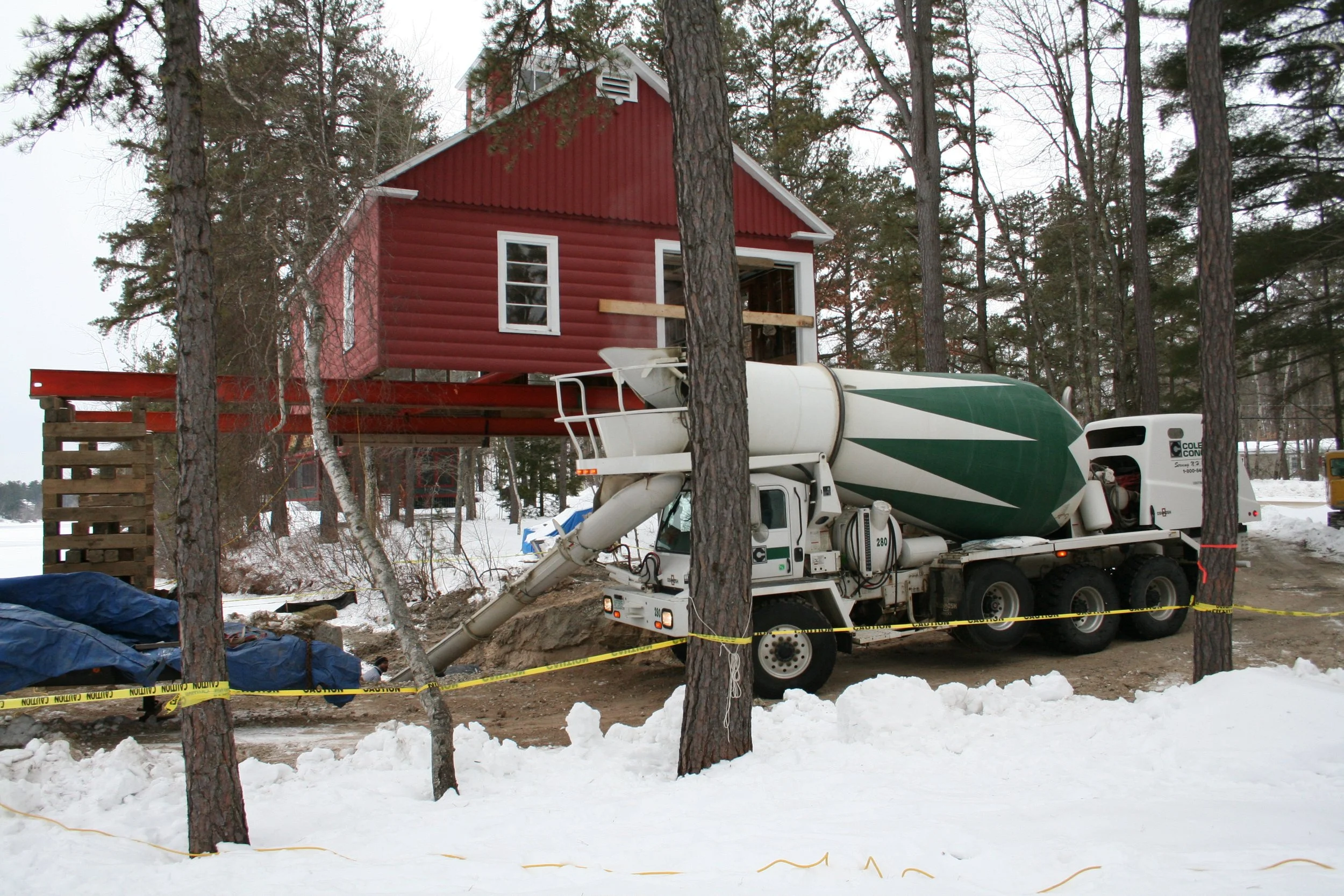 A construction site with a red house being built on stilts, with a cement mixer truck positioned in front, and snow on the ground surrounded by trees.
