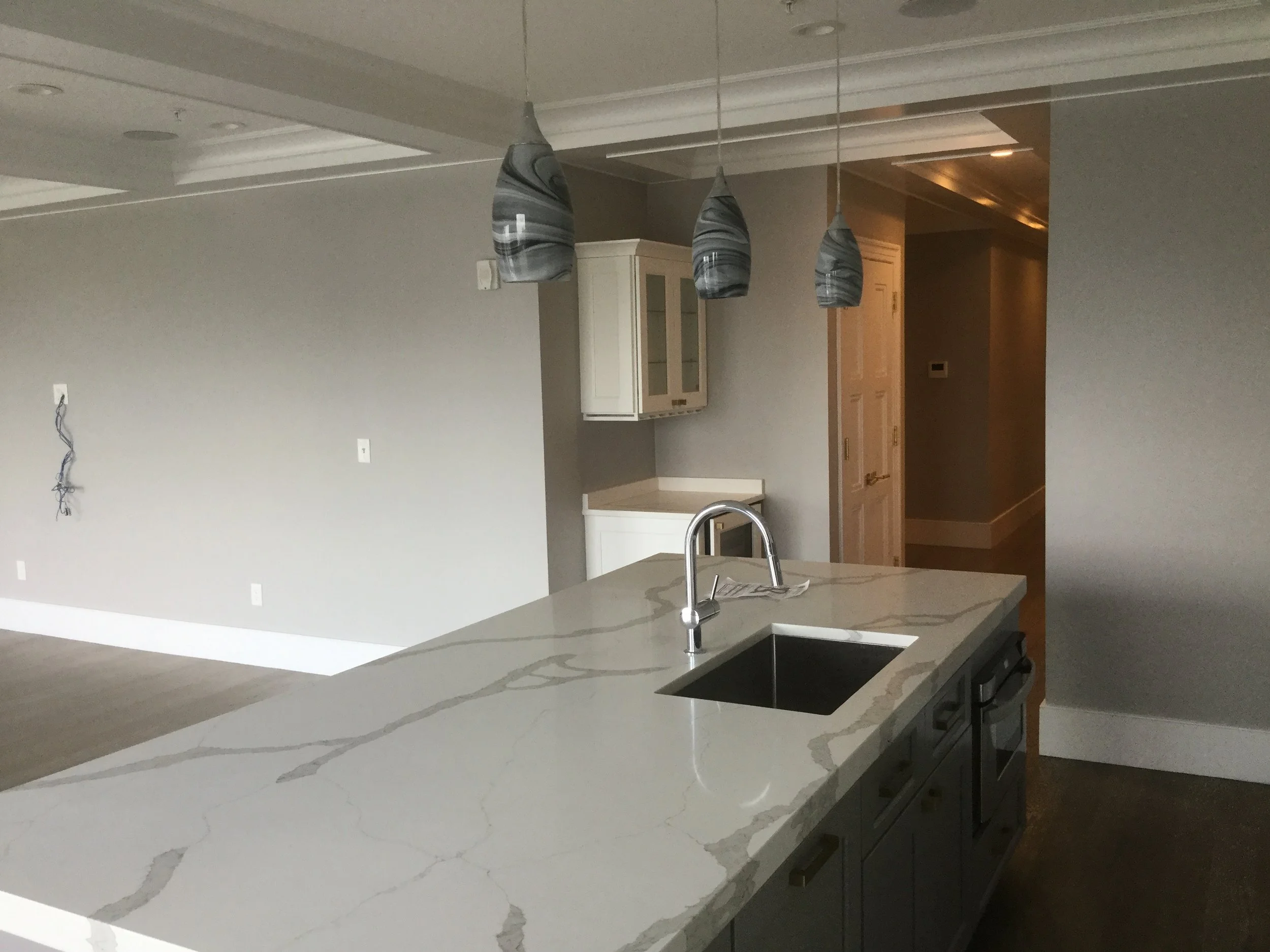 Empty modern kitchen with a white marble island counter, three gray and black pendant lights hanging above, gray cabinets, and a stainless steel sink.