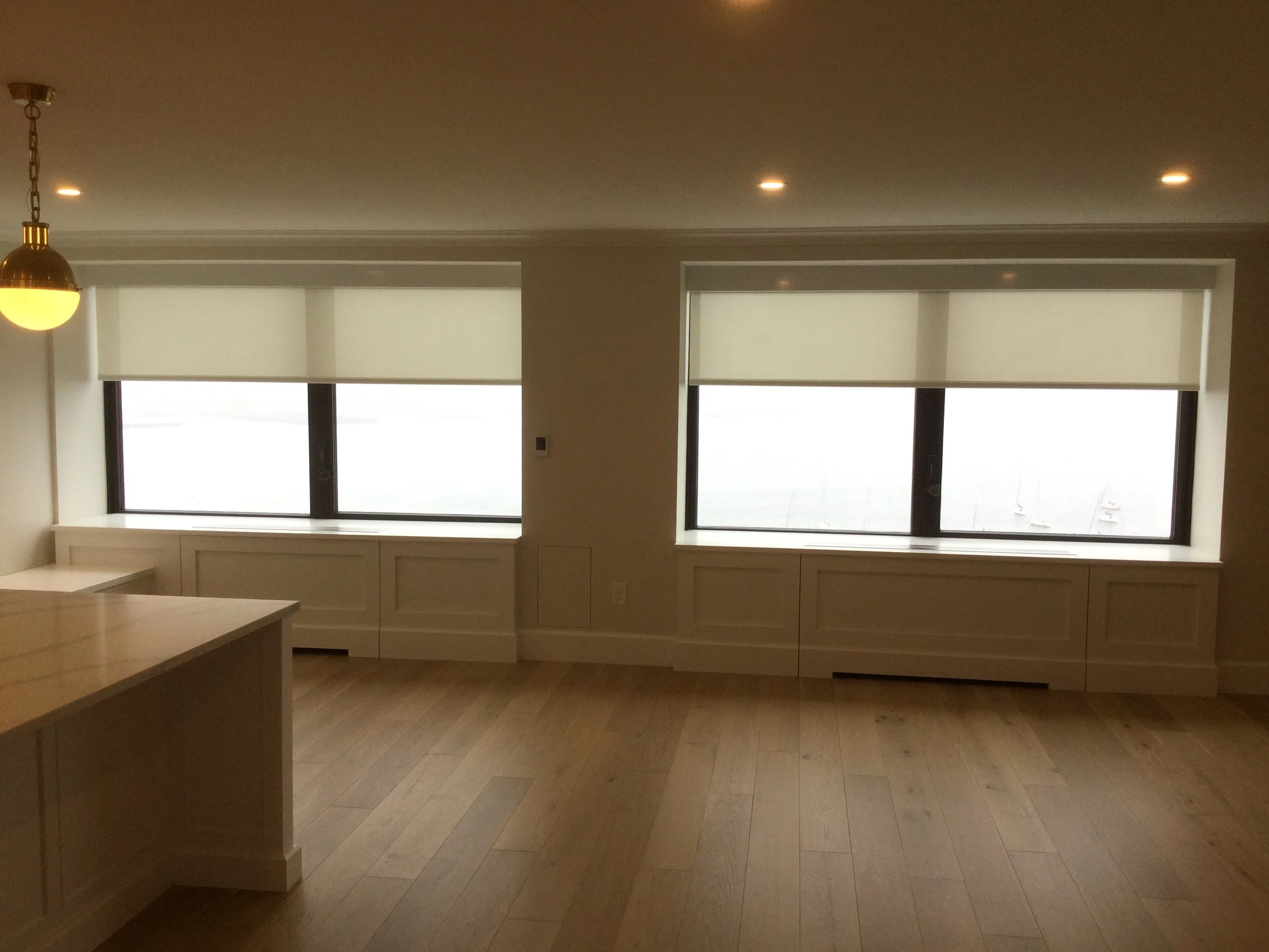An empty room with two large windows with white blinds, wooden floors, a corner of a kitchen island, and a ceiling light fixture, with a view of sailboats on the water outside.