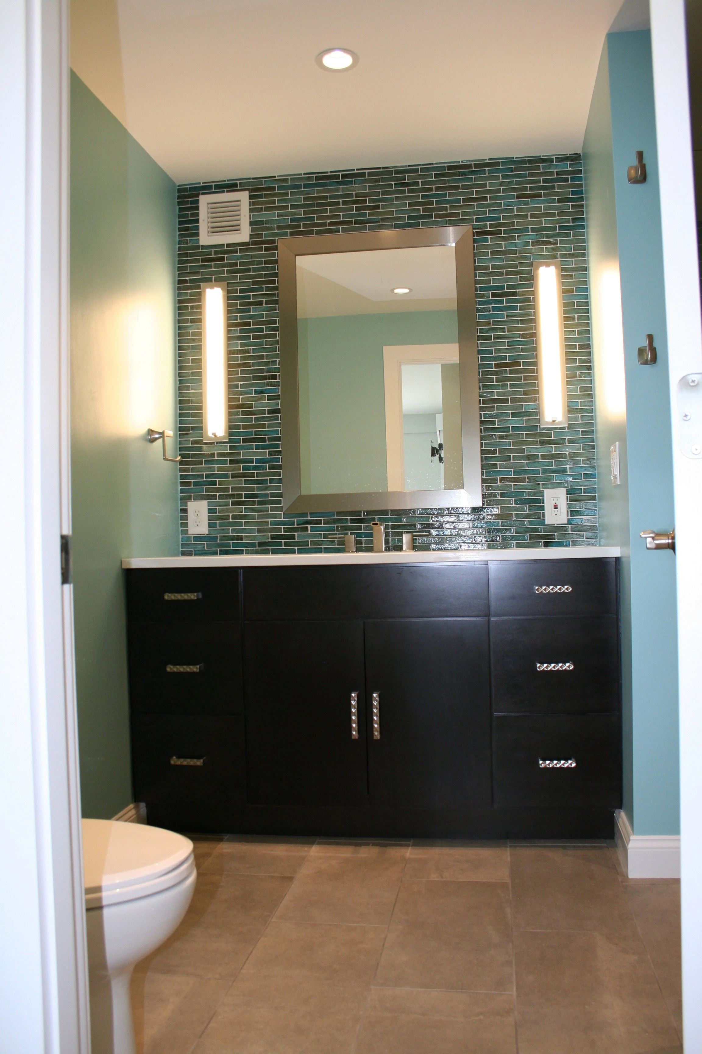 Modern bathroom with green and teal tiled accent wall, black vanity with silver handles, large mirror, vertical light fixtures, toilet in the foreground, and beige tiled floor.