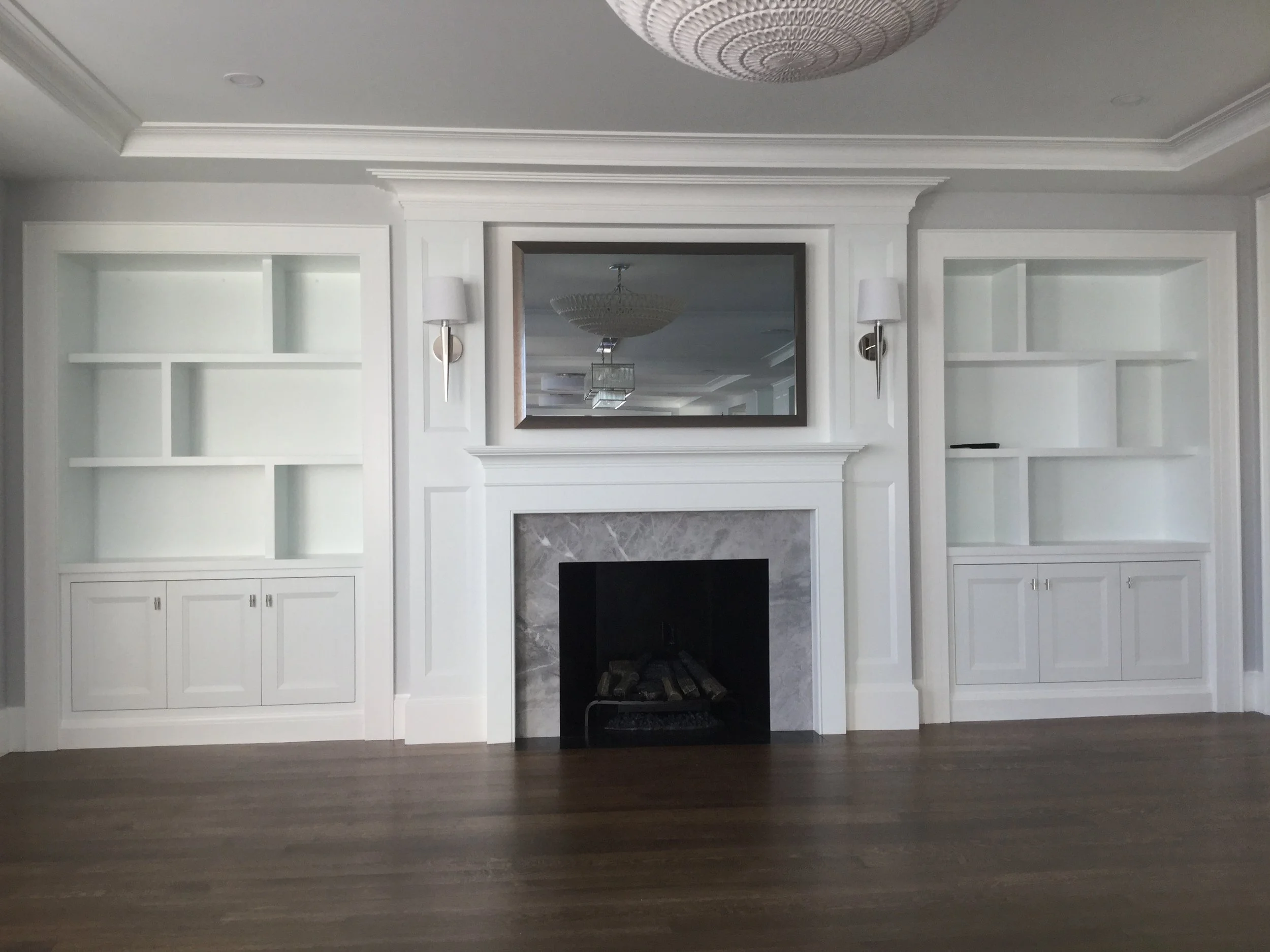 Living room with white built-in shelves on each side of a fireplace with a marble surround, a large mirror above the mantel, and wall sconces on either side of the mirror.