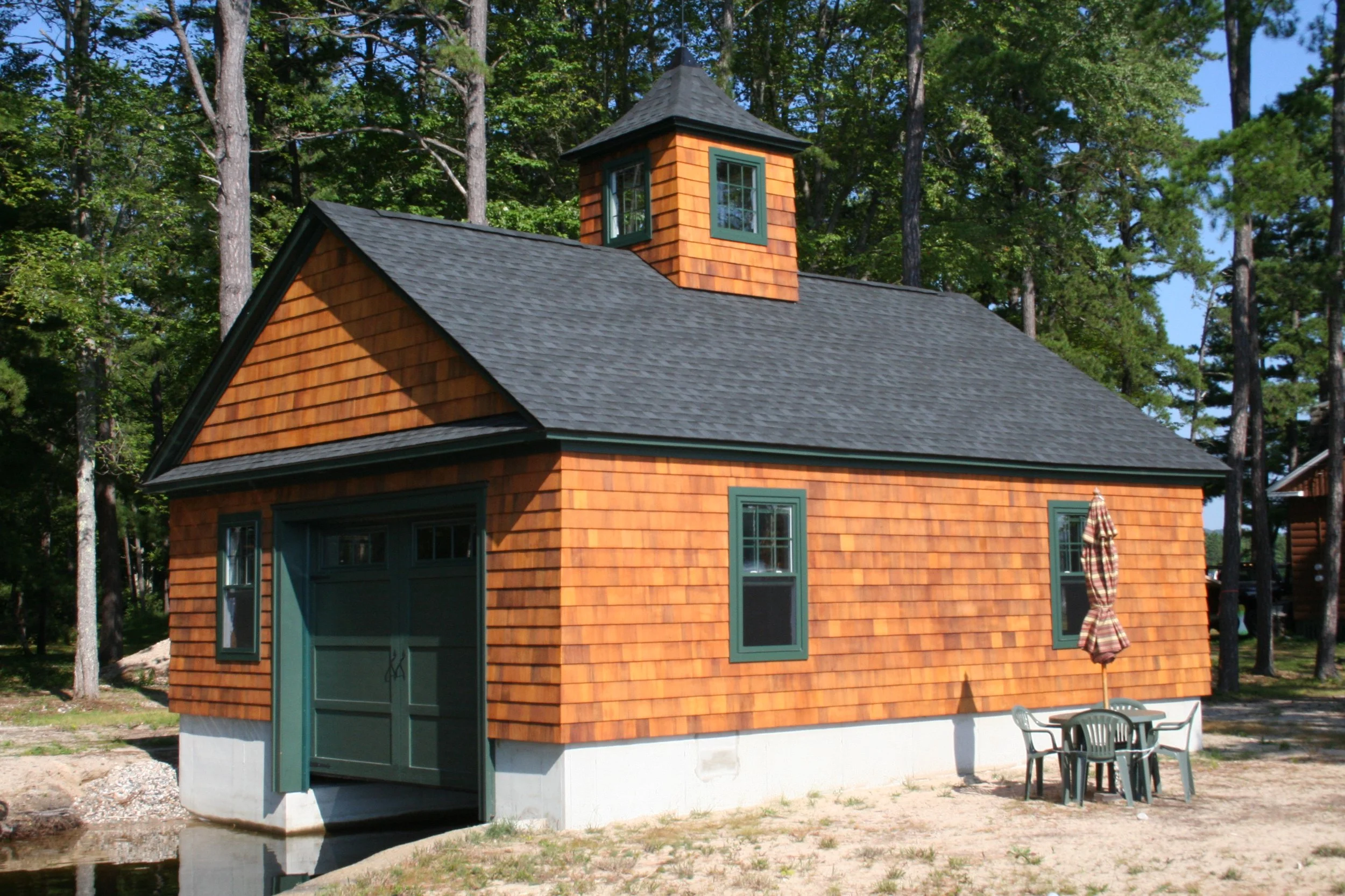 A small wooden building with a garage door has a black shingle roof and a small tower. There are four windows with green trim, and a table with four chairs and an umbrella outside.