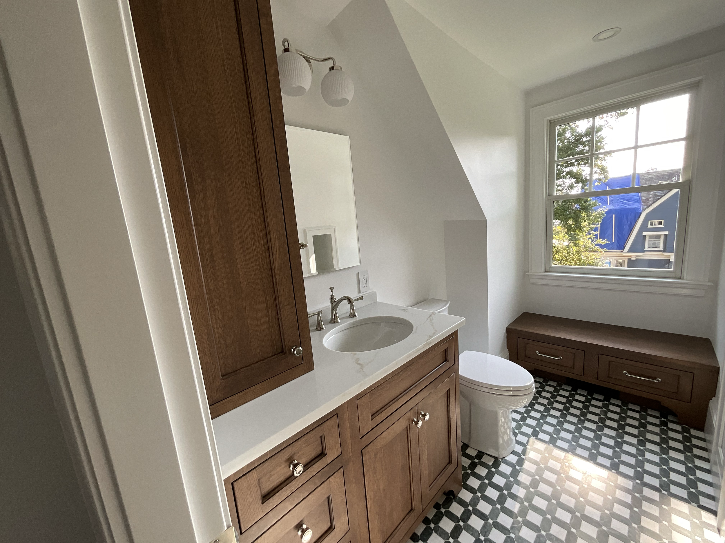 A bathroom with a wooden vanity, a marble countertop, a white toilet, a large window, and a bench with drawers. The floor has black and white patterned tile, and there is a mirror and two light fixtures above the sink.