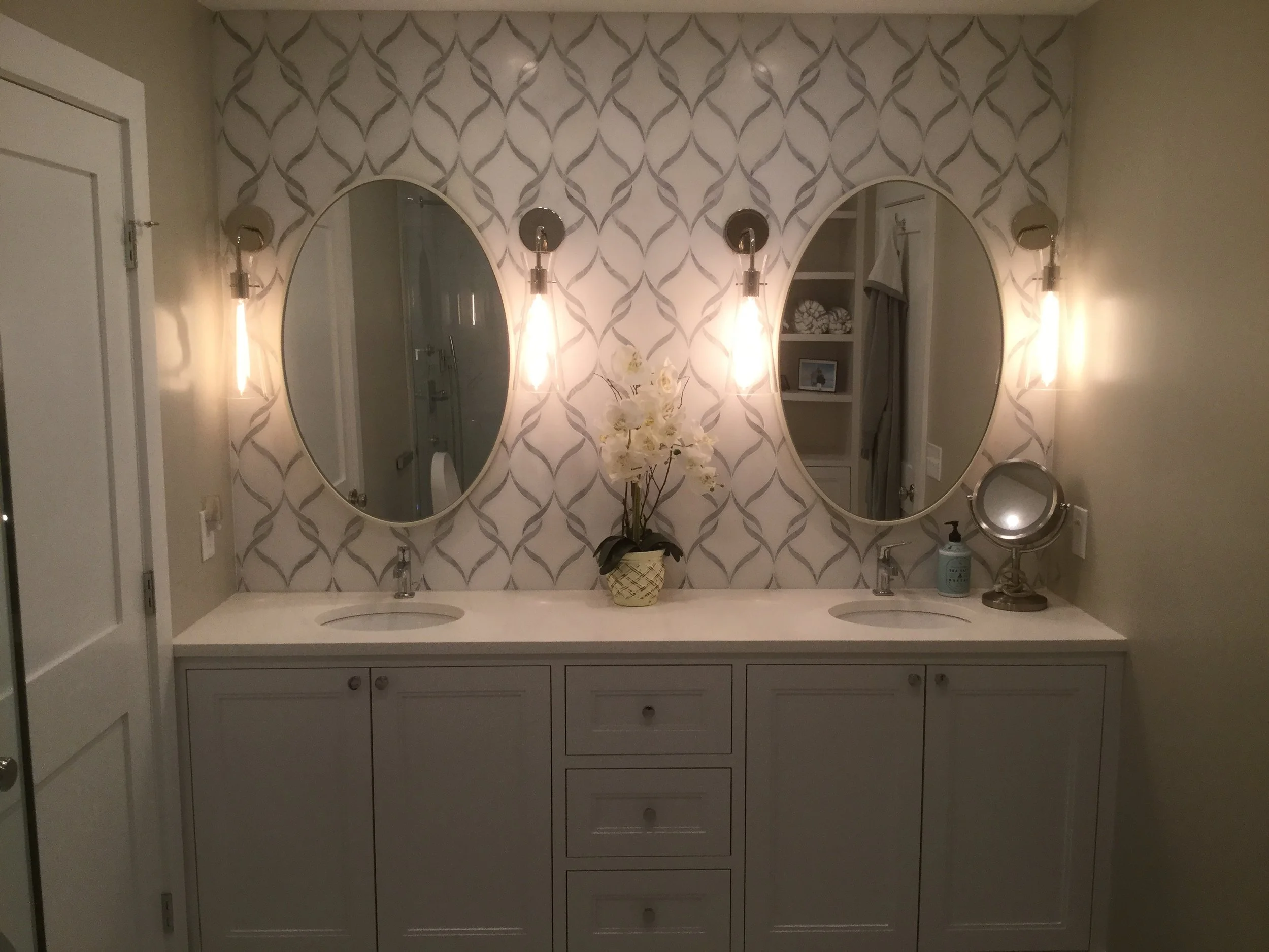 Double bathroom vanity with two oval mirrors, wall-mounted lights on each side, a flower arrangement in the middle, and various toiletries on the right side, with a patterned wallpaper background.