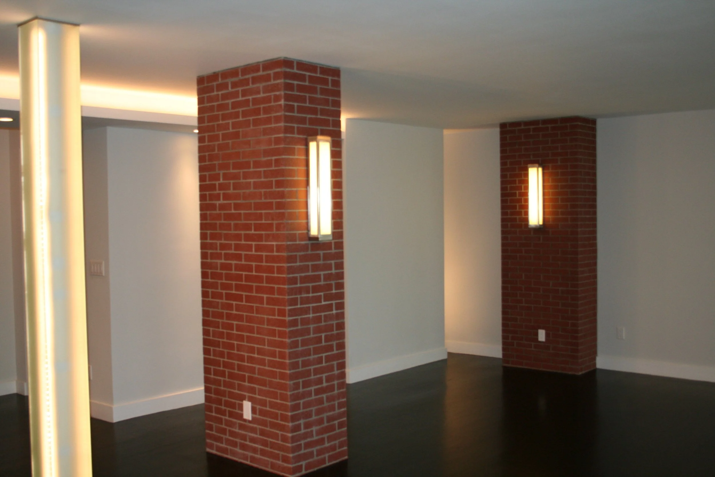 Interior of a modern room with two exposed red brick columns, white walls, dark wooden flooring, and vertical wall-mounted light fixtures.