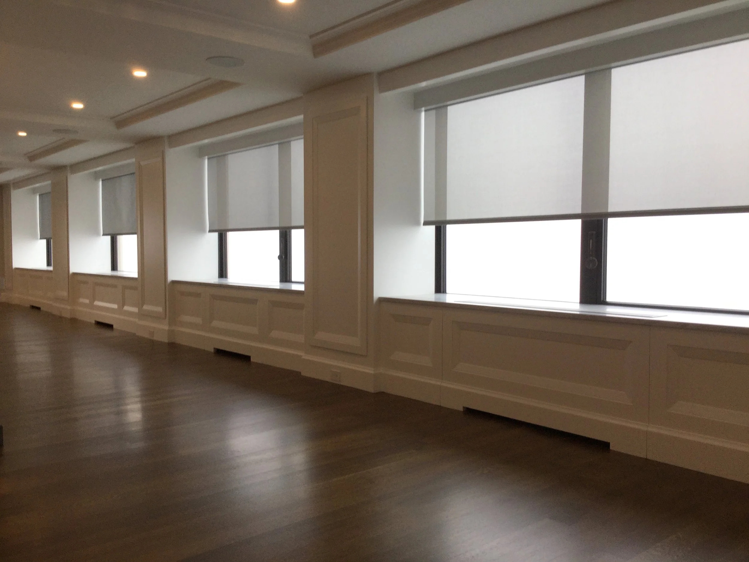Empty room with large windows and white window blinds, wooden flooring, and ceiling lights.