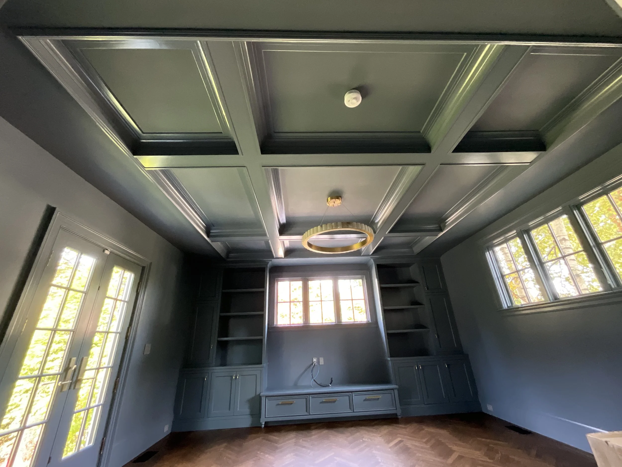 Empty blue room with built-in shelves, large windows, French doors, wooden floor, and modern gold chandelier.