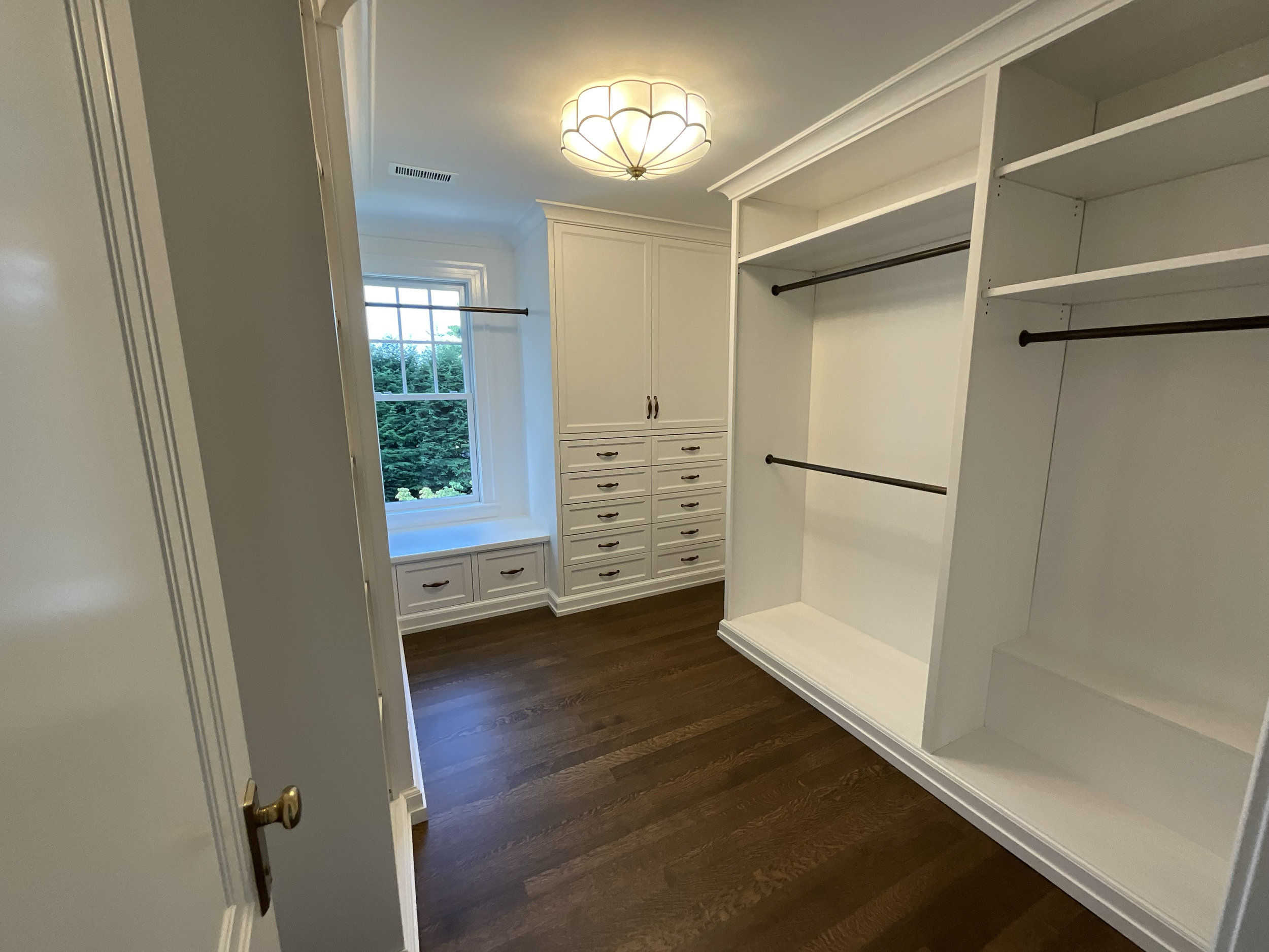 Empty walk-in closet with white built-in shelving and drawers, dark wood flooring, a window with a seat, and a ceiling light fixture.