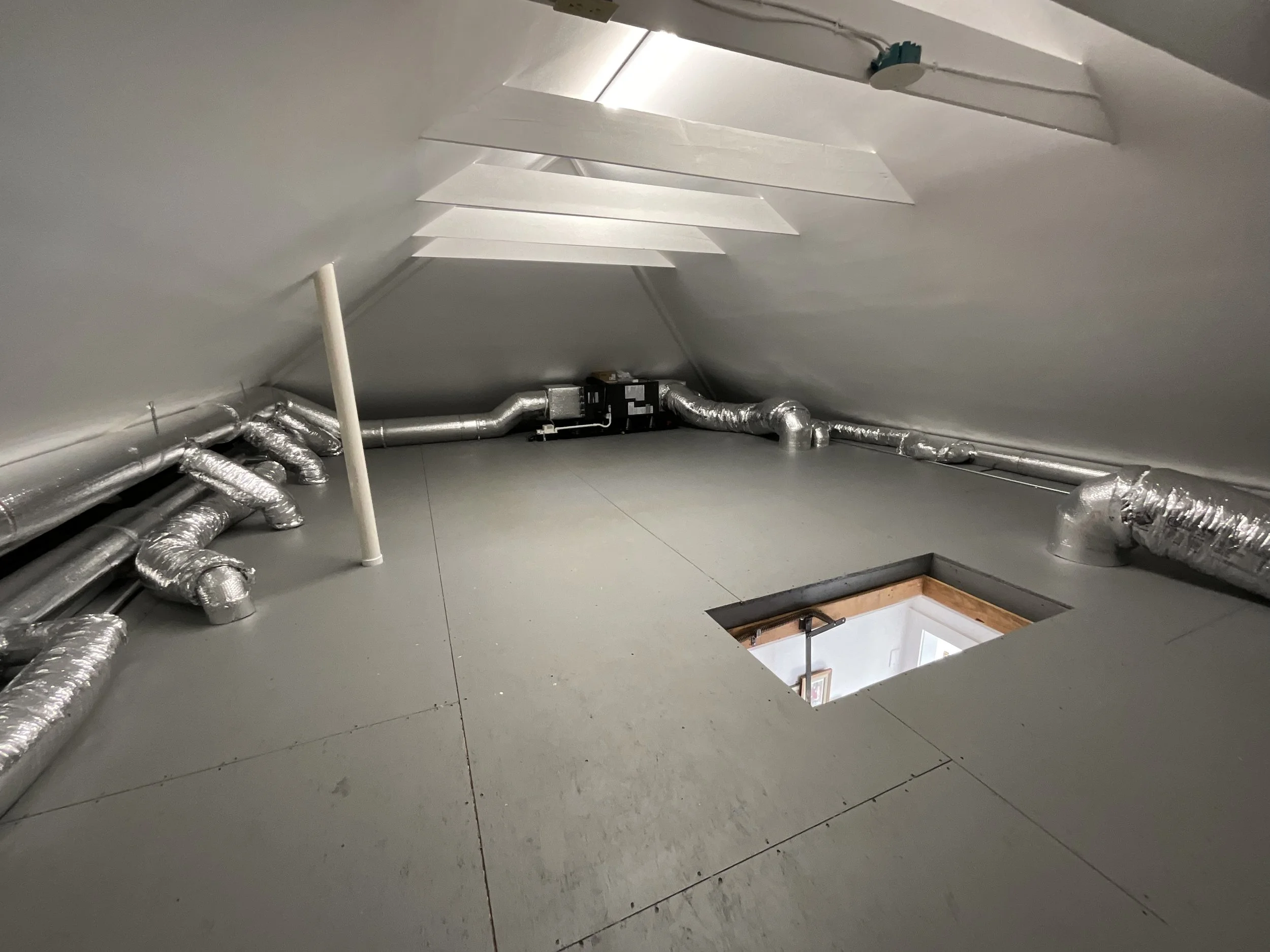 Attic space with silver insulated ductwork running along the floor and walls. There is a small square opening in the floor and ceiling with a wooden frame around it.