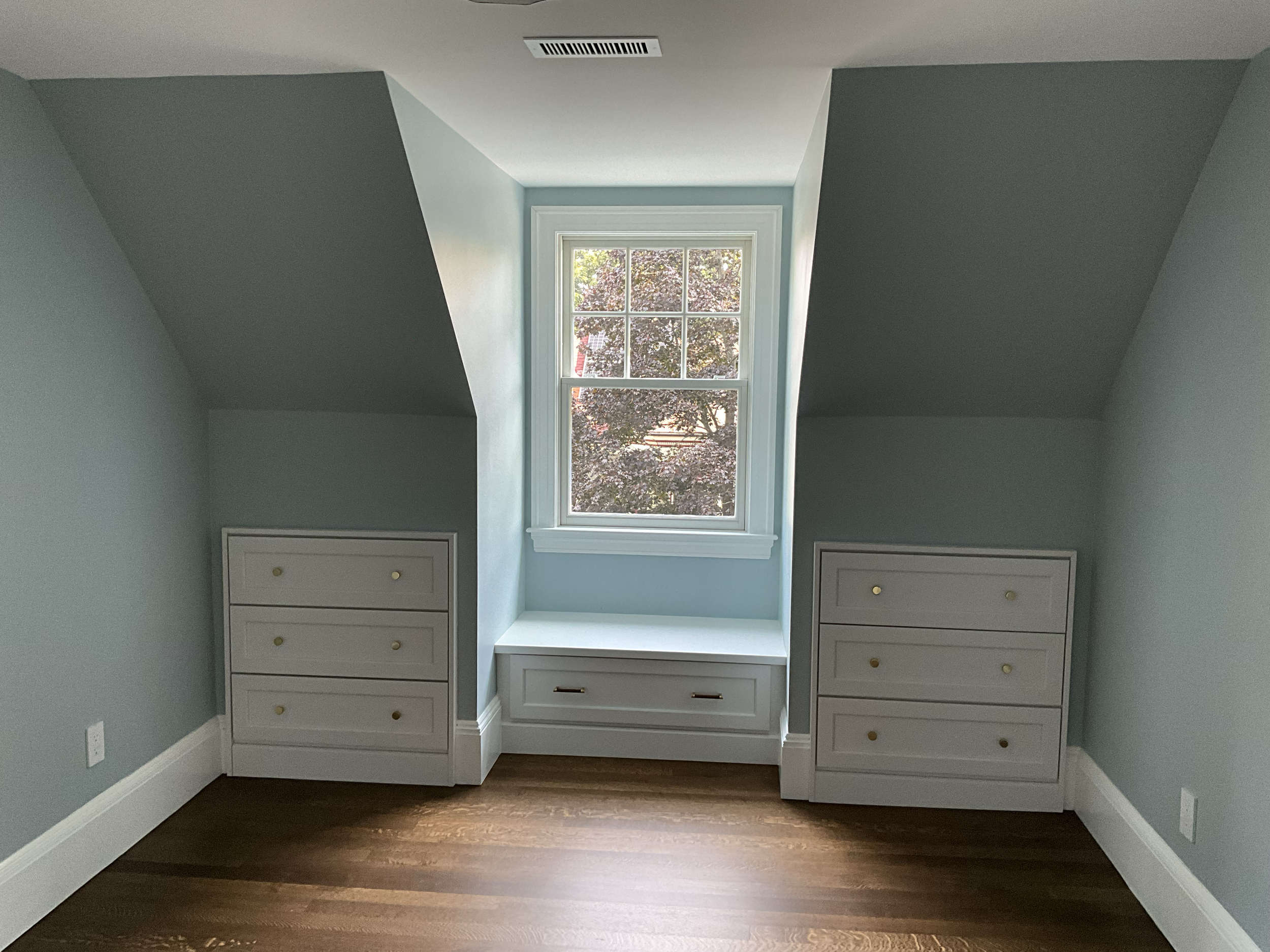 A small, empty bedroom with light blue walls, a white window with a view of trees outside, two white dressers with gold knobs, and a wooden floor.