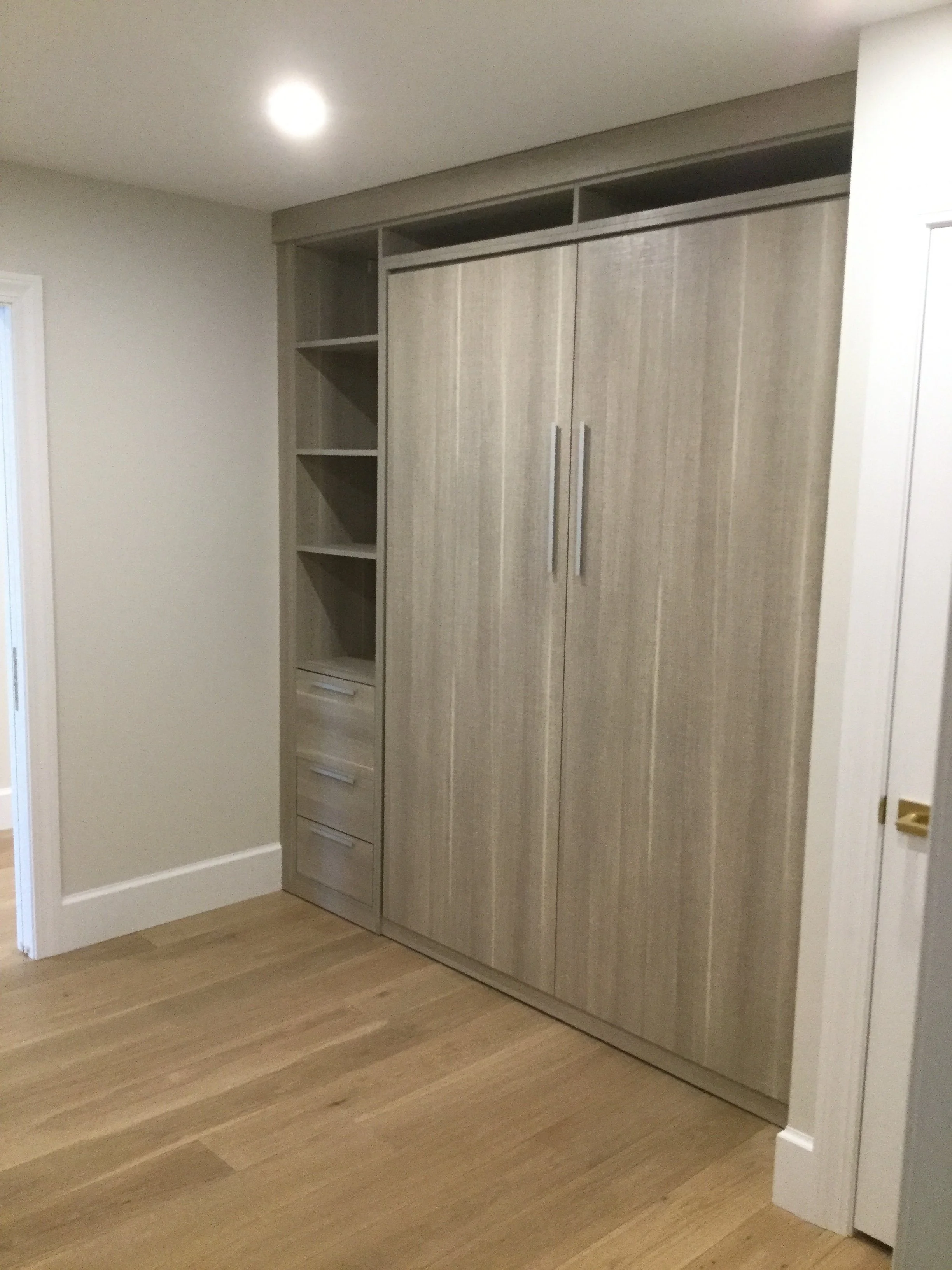 A built-in closet with closed double doors, open shelves on the side, and drawers at the bottom, in a room with wood flooring and a white wall.