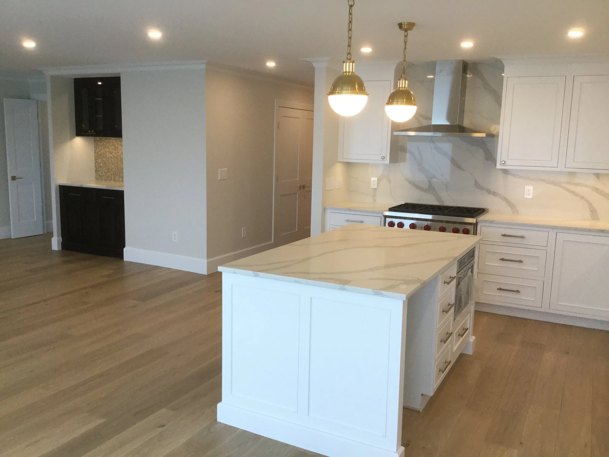 Modern kitchen with white cabinets, marble countertops, wooden flooring, and gold pendant lights.