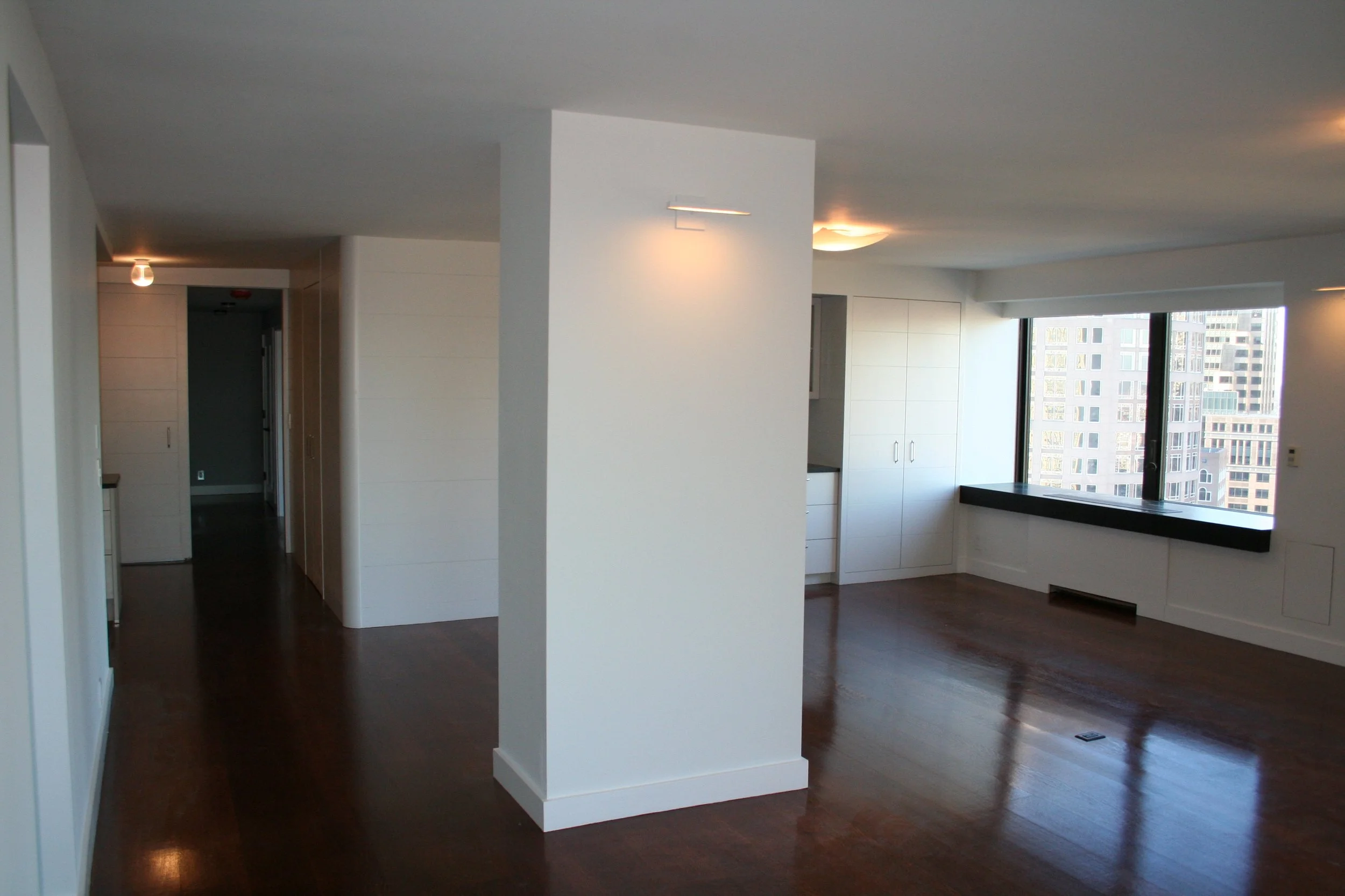 Empty living room with large windows, white walls, dark hardwood floors, built-in white cabinetry, and cityscape view.