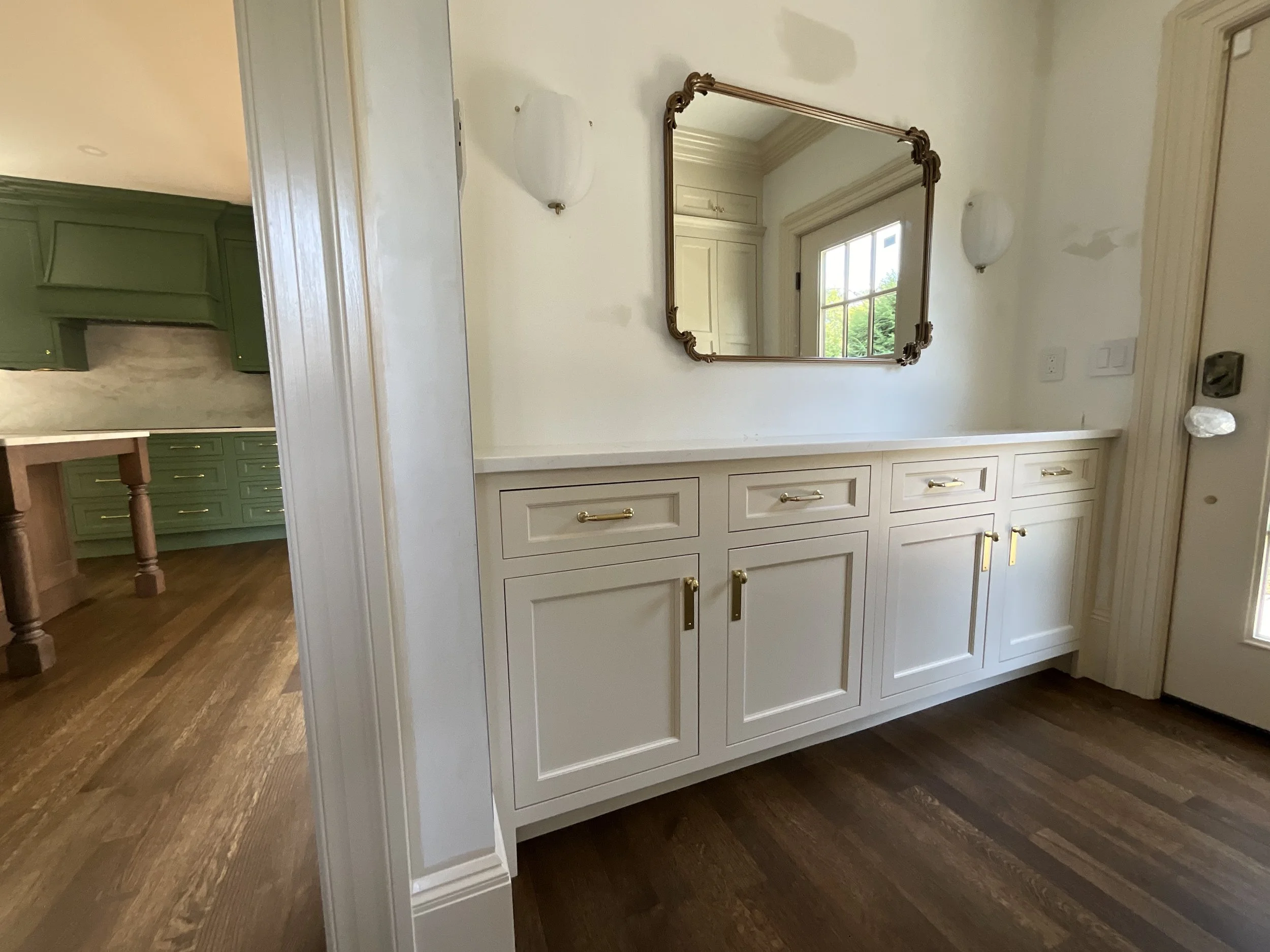 Interior view of a white built-in cabinet with gold handles, a wall mirror, and wall-mounted lights in a home, with a kitchen in the background featuring green cabinetry and wooden flooring.