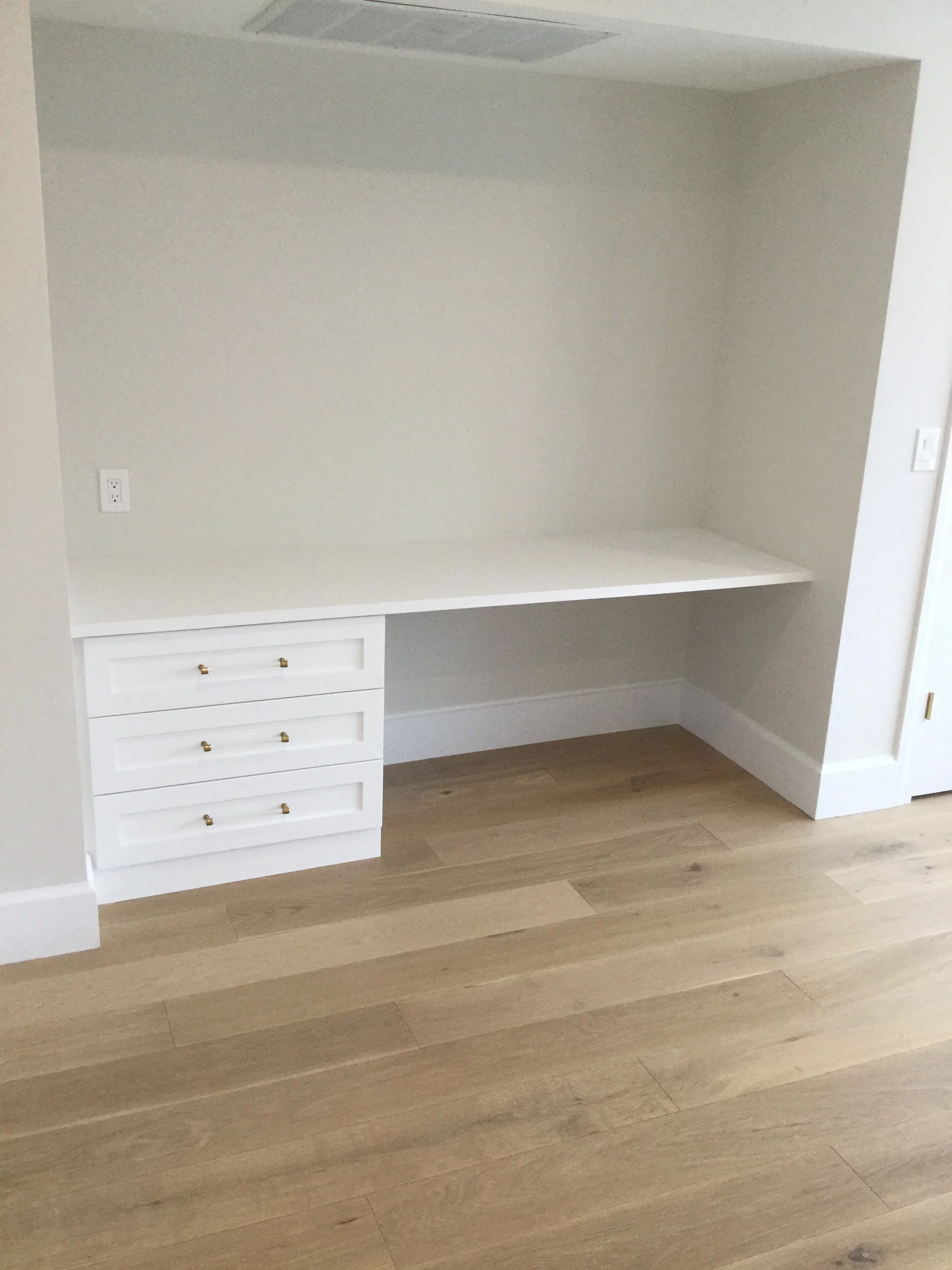 Empty built-in desk or workspace with a white countertop and three drawers beneath, set against a wall with electrical outlets, next to a doorway.