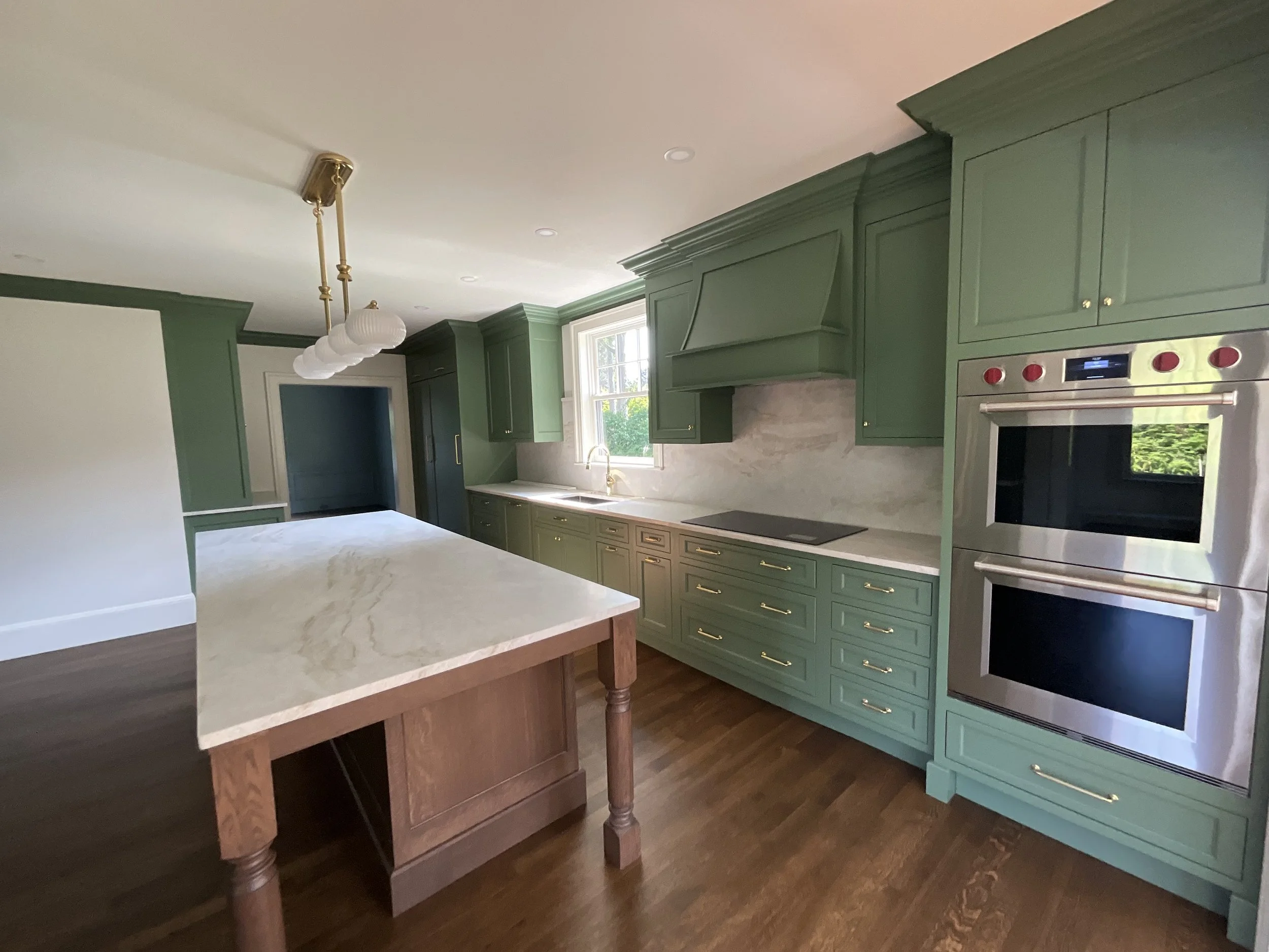 Modern kitchen with green cabinets, gold hardware, marble countertops, double oven, and wooden island.
