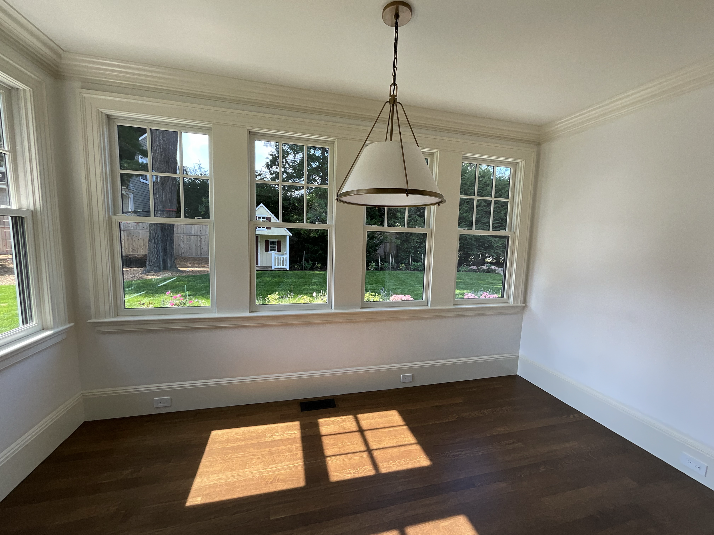 Empty room with hardwood floors, large windows with white trim, and a white hanging light fixture. Outside the windows, a yard with green grass, a tree, garden, and a small playhouse are visible.