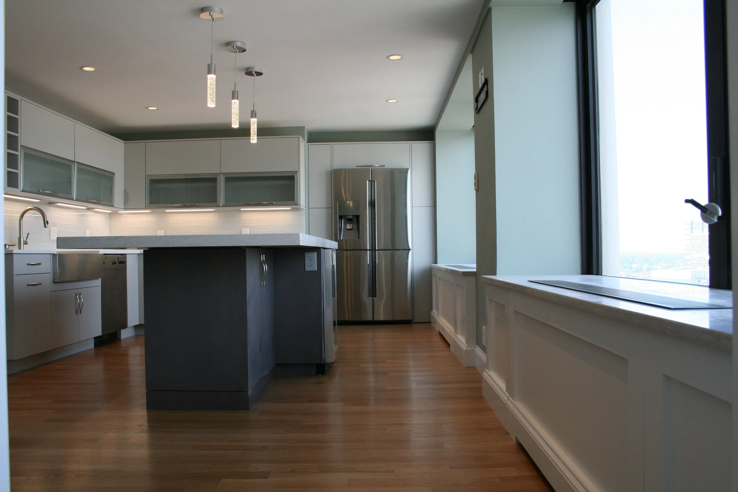 Modern kitchen with white cabinets, stainless steel refrigerator, island with gray base, and wood flooring, overlooking cityscape through large windows.