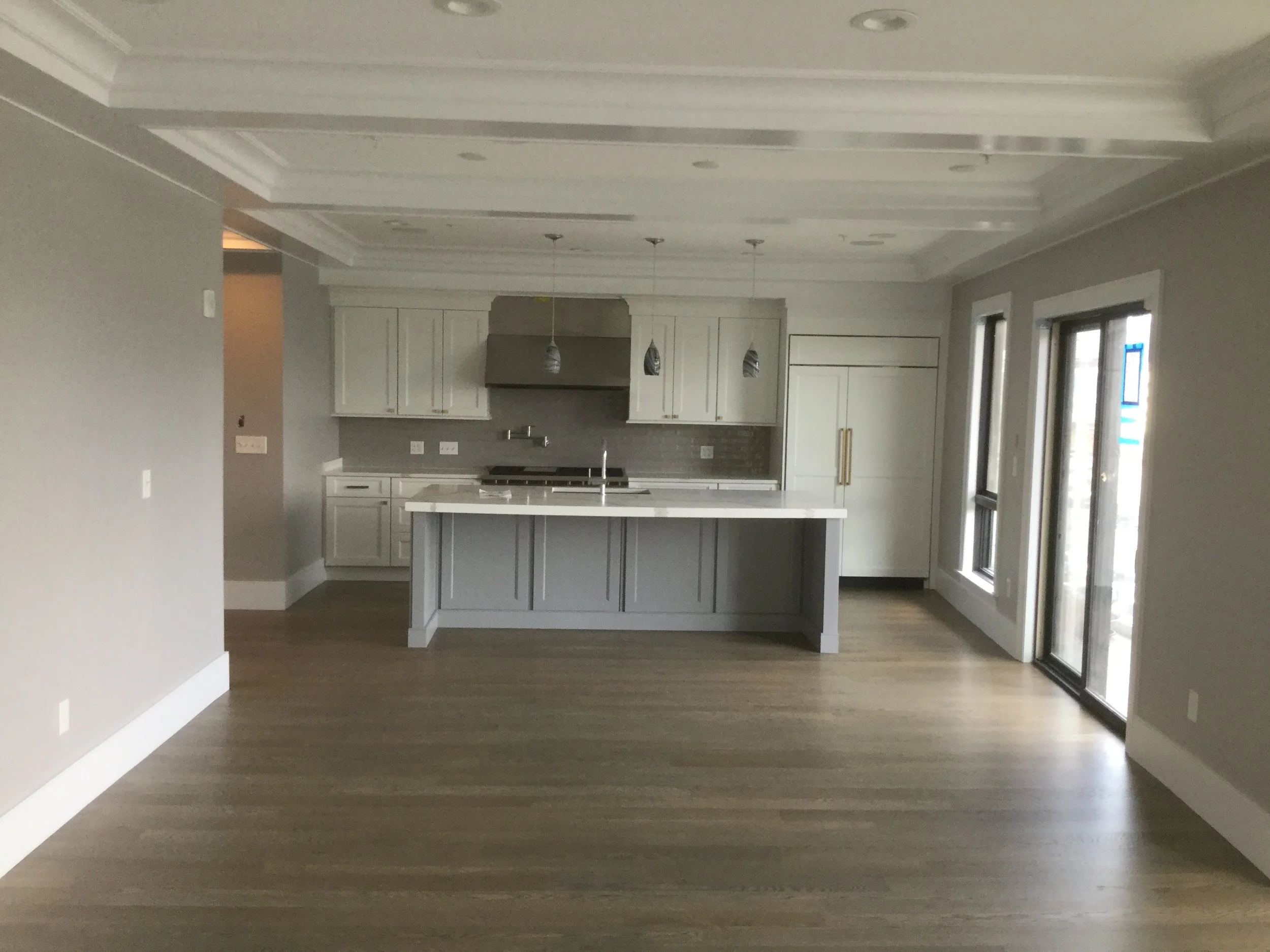 Empty modern kitchen with white cabinets, a kitchen island, stainless steel appliances, and large windows letting in natural light.