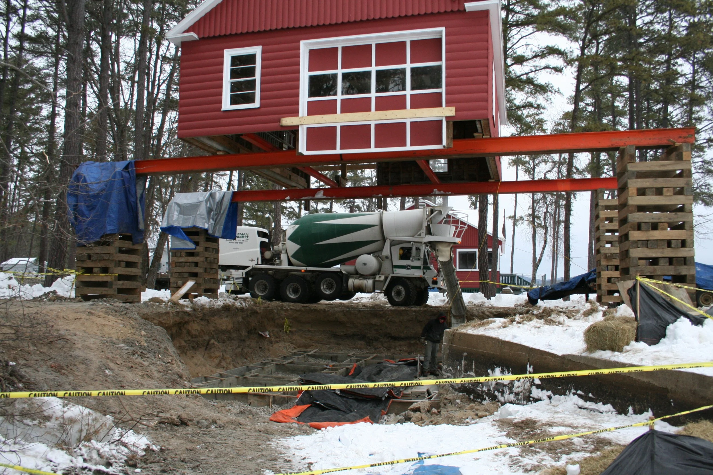 Construction site with a red house elevated on stilts, a cement mixer truck pouring concrete into a foundation, and workers on snowy ground.