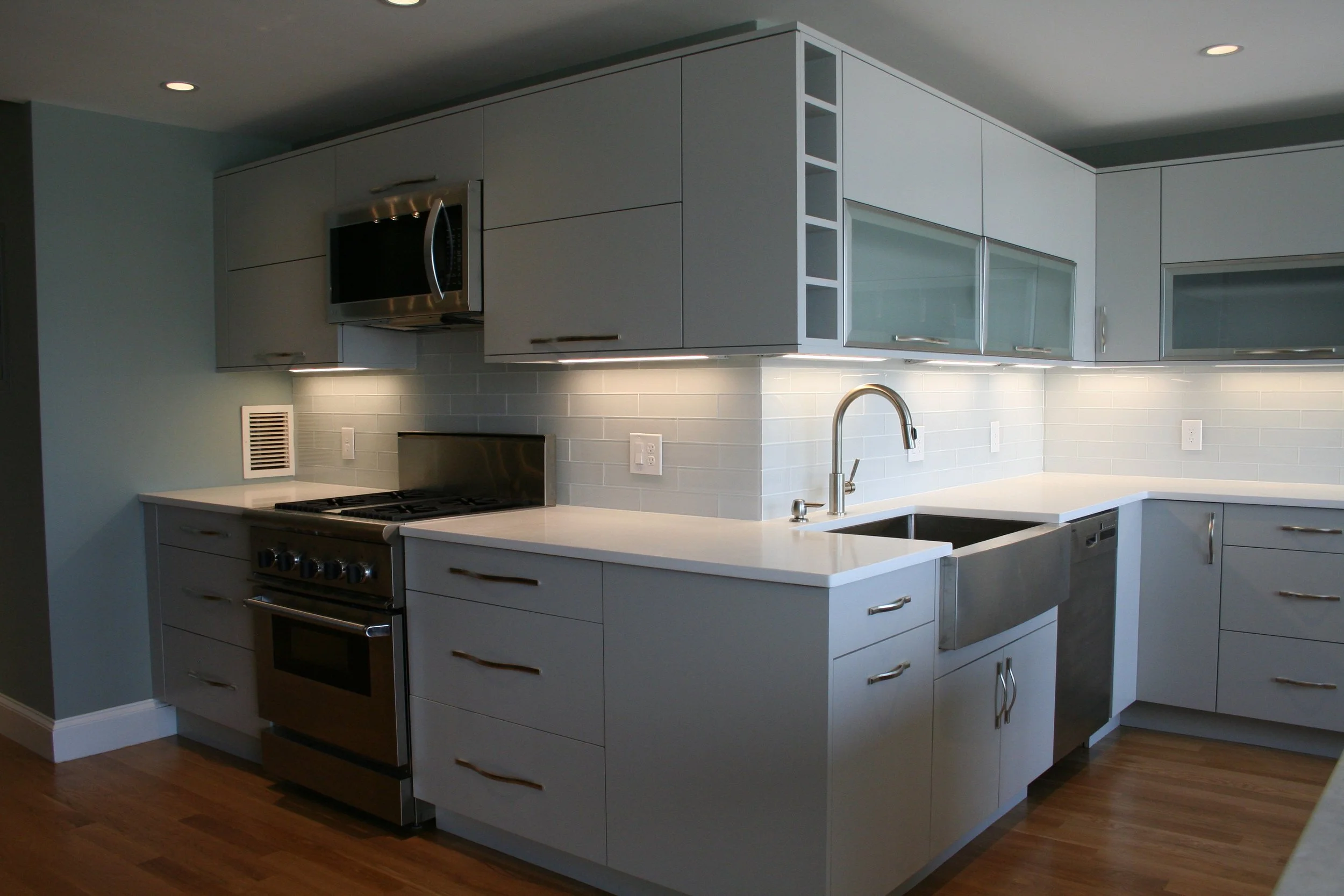 Modern kitchen with white countertops, gray cabinets, stainless steel appliances, and wooden flooring.