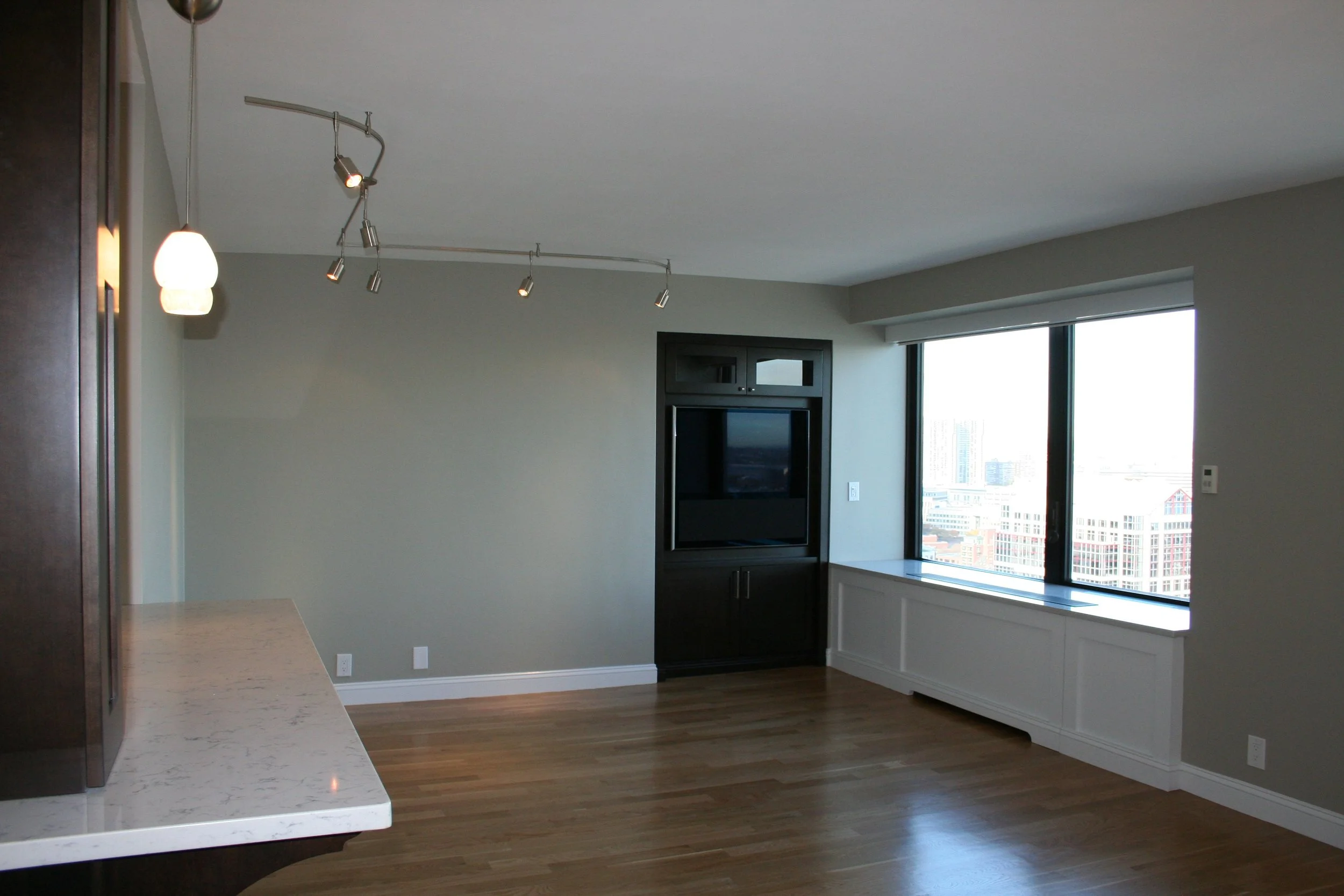 Empty living room with large window, wooden floor, and a built-in TV unit, with a partial view of a kitchen counter in the foreground.