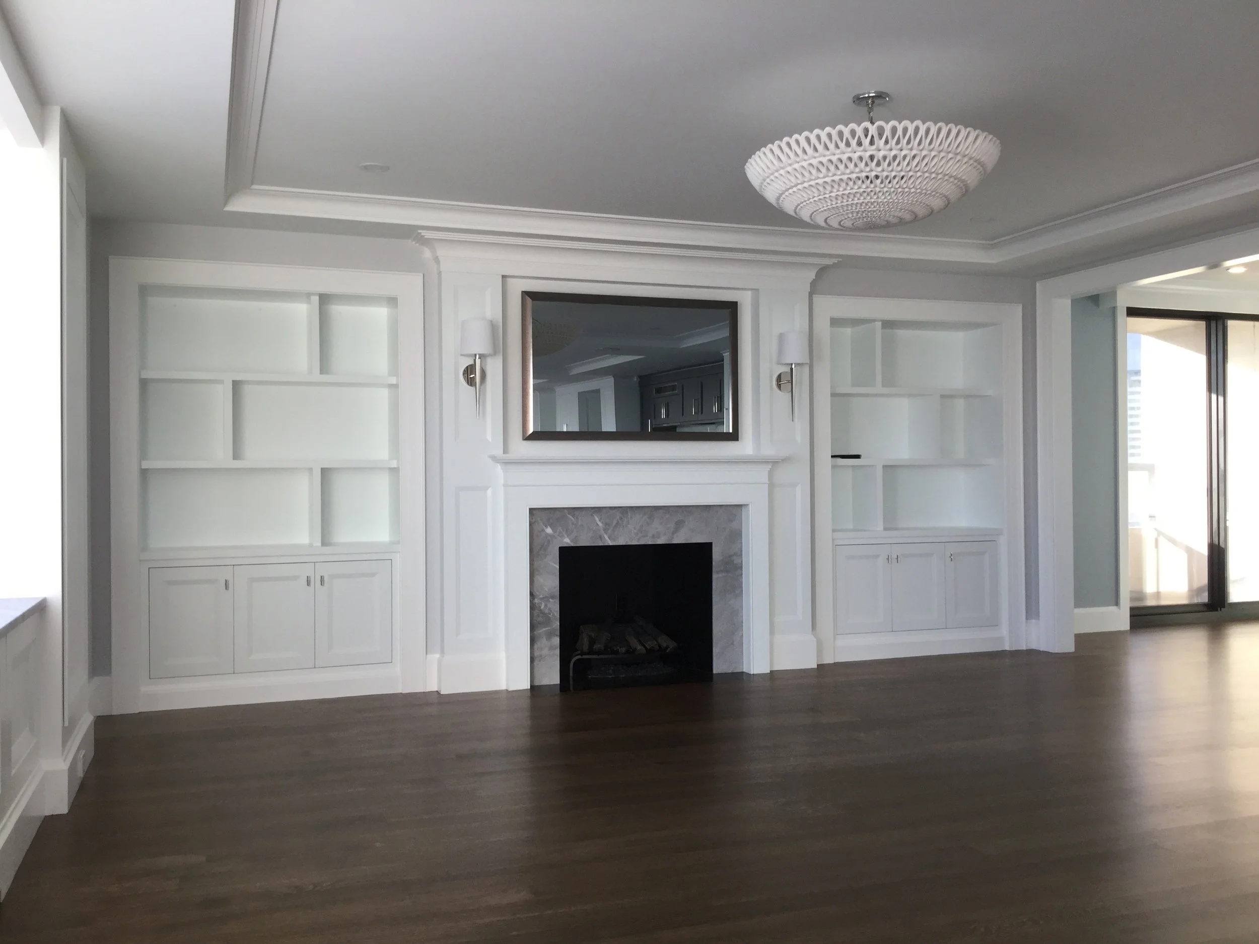 Empty living room with white built-in shelves, fireplace with gray marble surround, mirror above, hardwood floors, and a sliding glass door leading to a balcony.