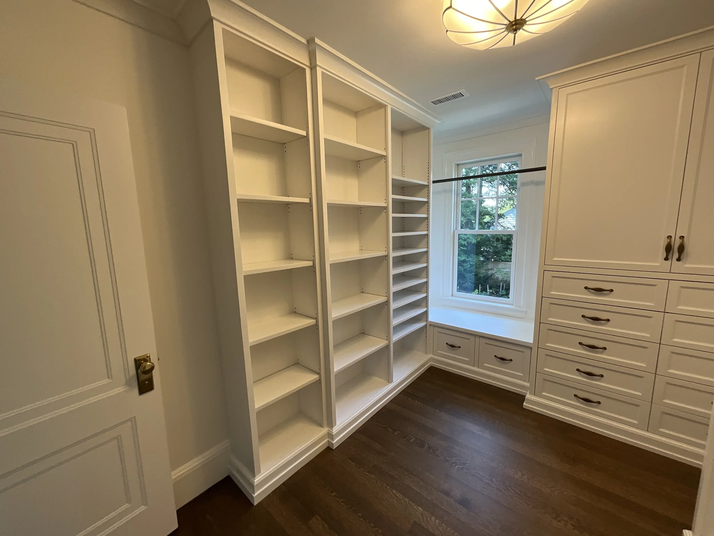 Empty walk-in closet with white built-in shelves, drawers, and cabinets, a large window, hardwood flooring, and a ceiling light fixture.