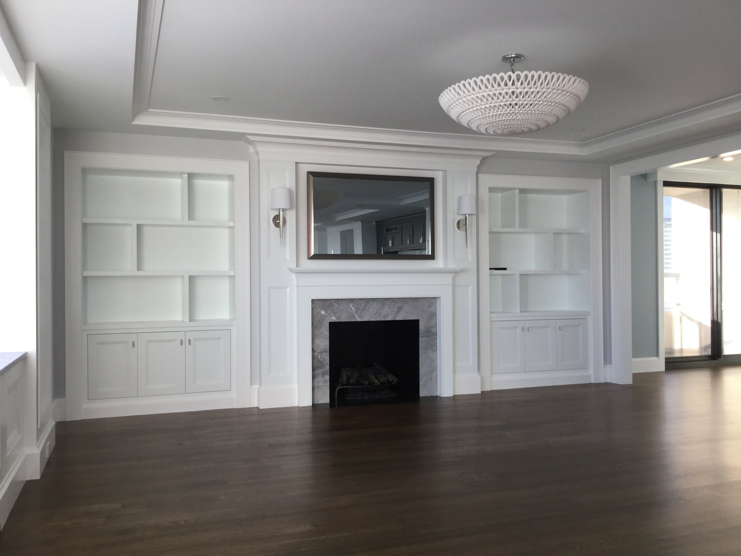 Living room with white built-in shelves on either side of a fireplace with a marble surround, a mirror above, wall sconces, a ceiling light fixture, wood flooring, and a sliding glass door leading to a balcony.