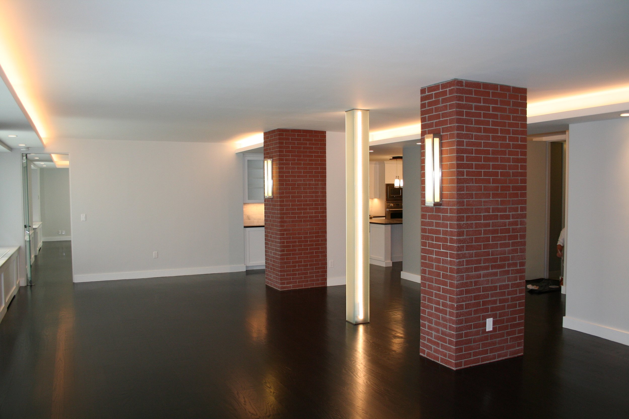 Empty modern living room with dark hardwood floors, white walls, and brick columns, lit by wall sconces and ceiling lights.