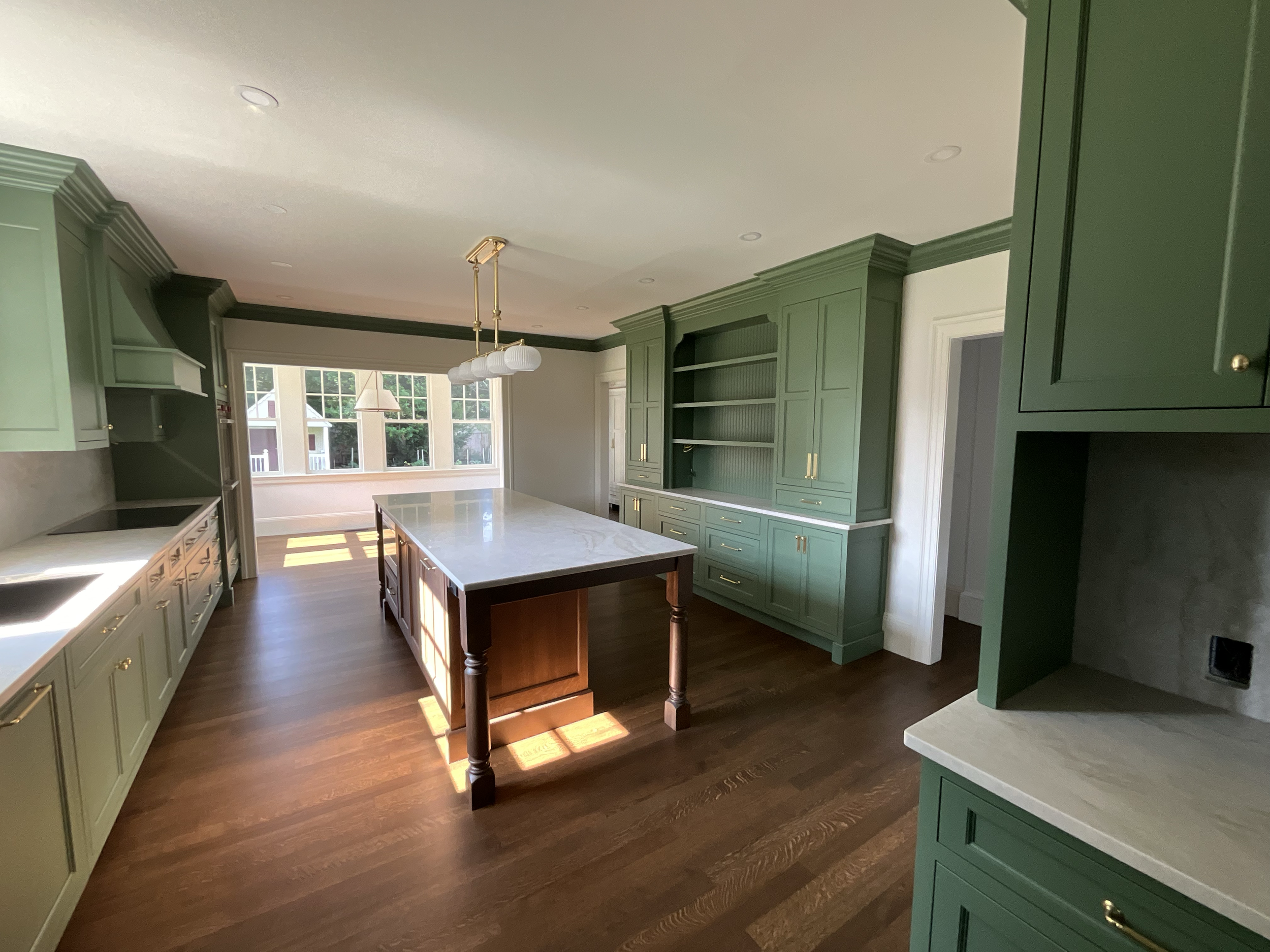 A spacious kitchen with green cabinets, a central island with a white marble top, hardwood floors, and large windows at the far end letting in natural light.