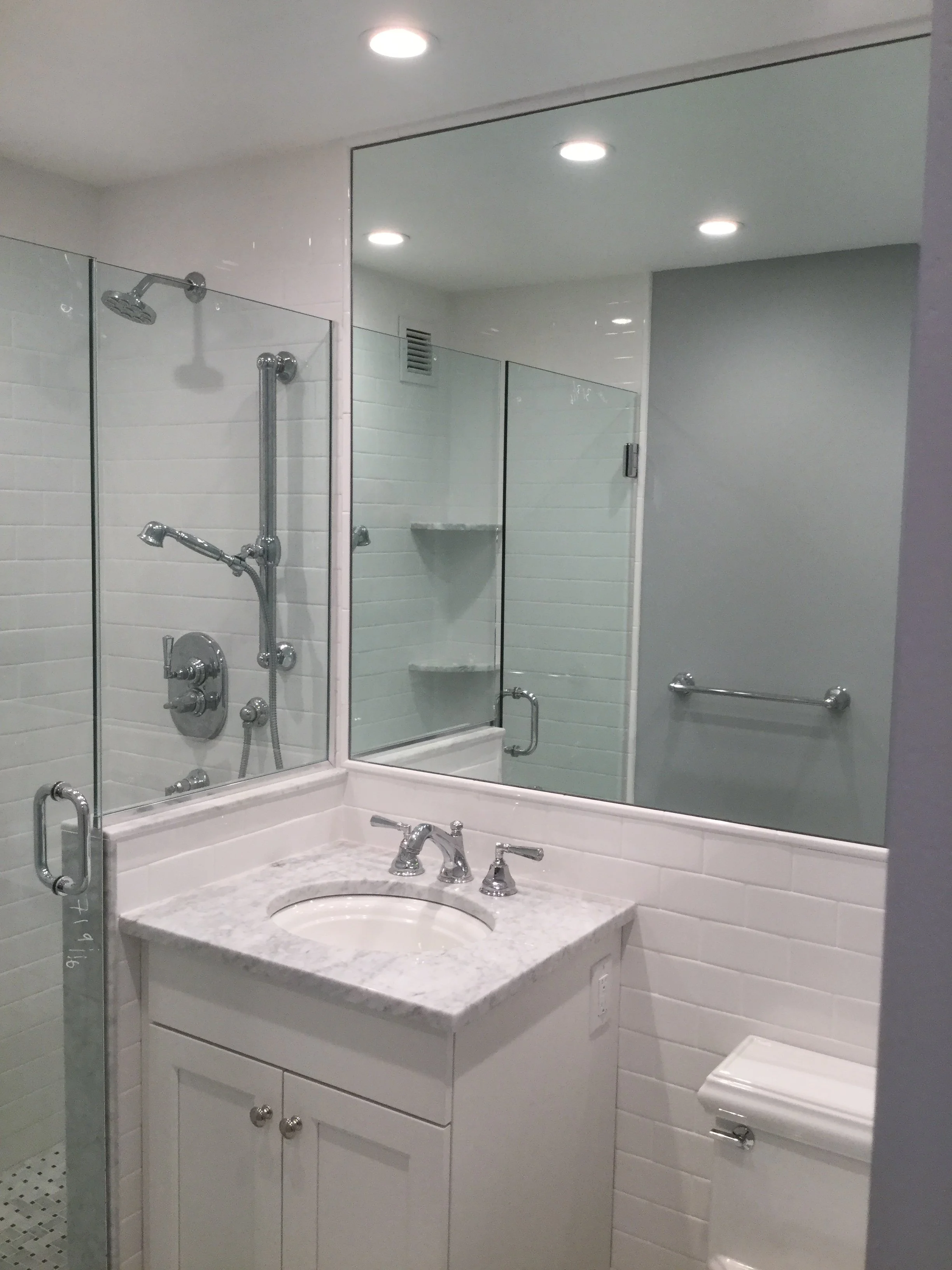 A clean bathroom with a shower enclosure, a mirror, a sink with a granite countertop, and a toilet. The bathroom has white tiled walls and recessed ceiling lights.