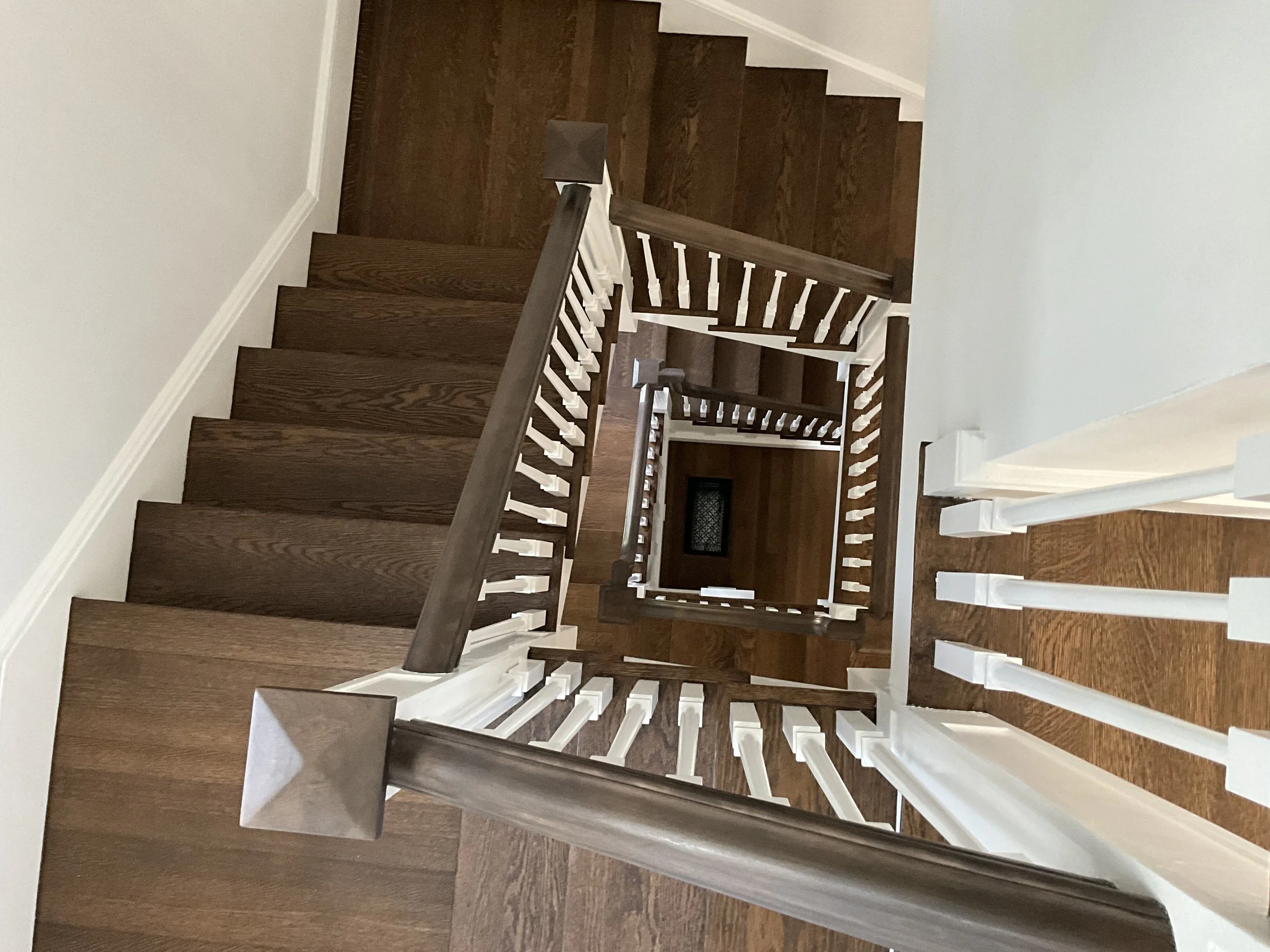 From the top view, this photo shows a wooden staircase with a dark brown handrail and white balusters, spiraling downward through several levels inside a house with tan floorboards.