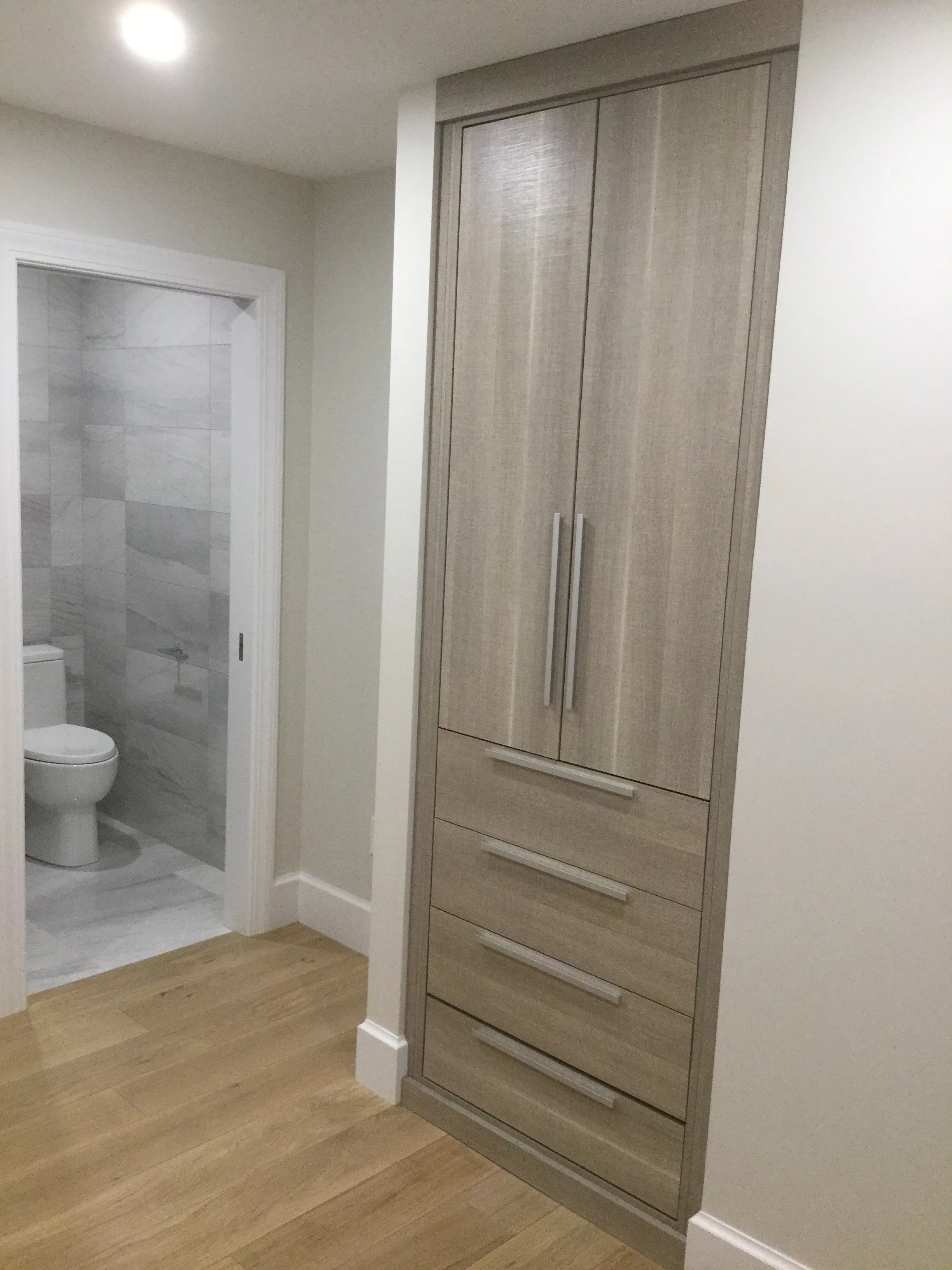 A tall wooden cabinet with multiple handles is placed in a corner near a doorway to a bathroom with a toilet and gray marble walls.