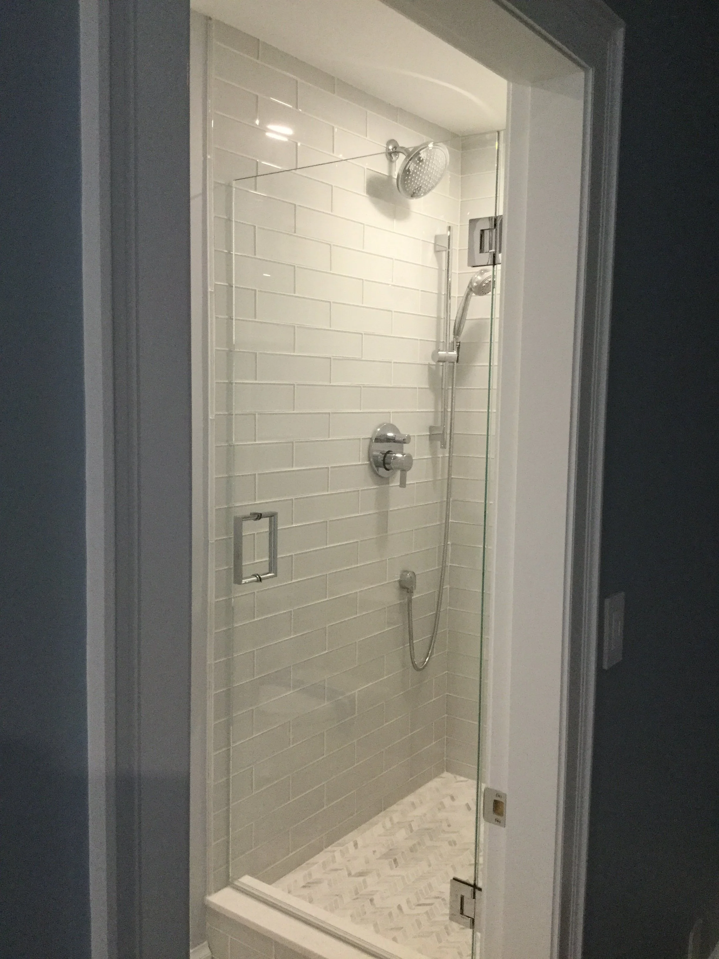 A modern shower stall with glass door, white subway tile walls, and a patterned tile floor, featuring a rain showerhead and handheld shower.